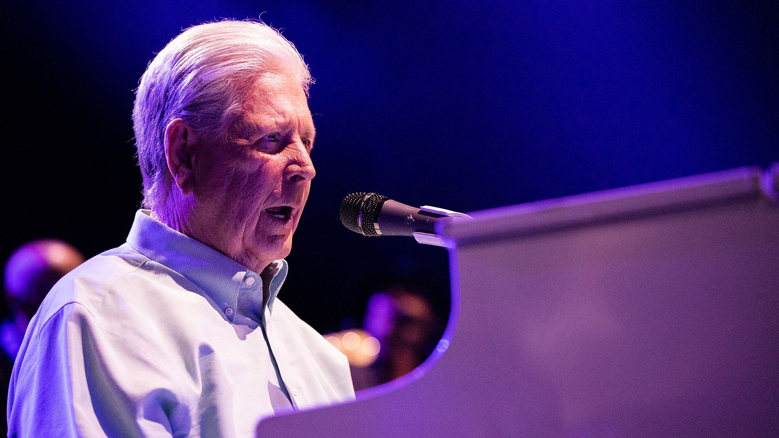 Brian Wilson, sentado al piano, canta al micrófono en un concierto con iluminación azul y violeta. Lleva una camisa de manga larga clara.