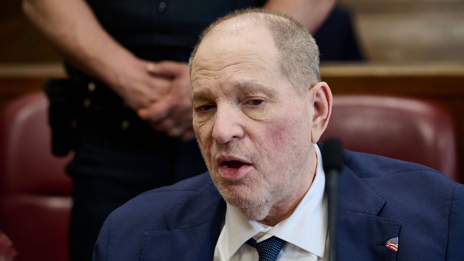 Imagen de Harvey Weinstein en su juicio en Nueva York, con traje azul marino y expresión seria, hablando. Un policía está detrás, desenfocado.
