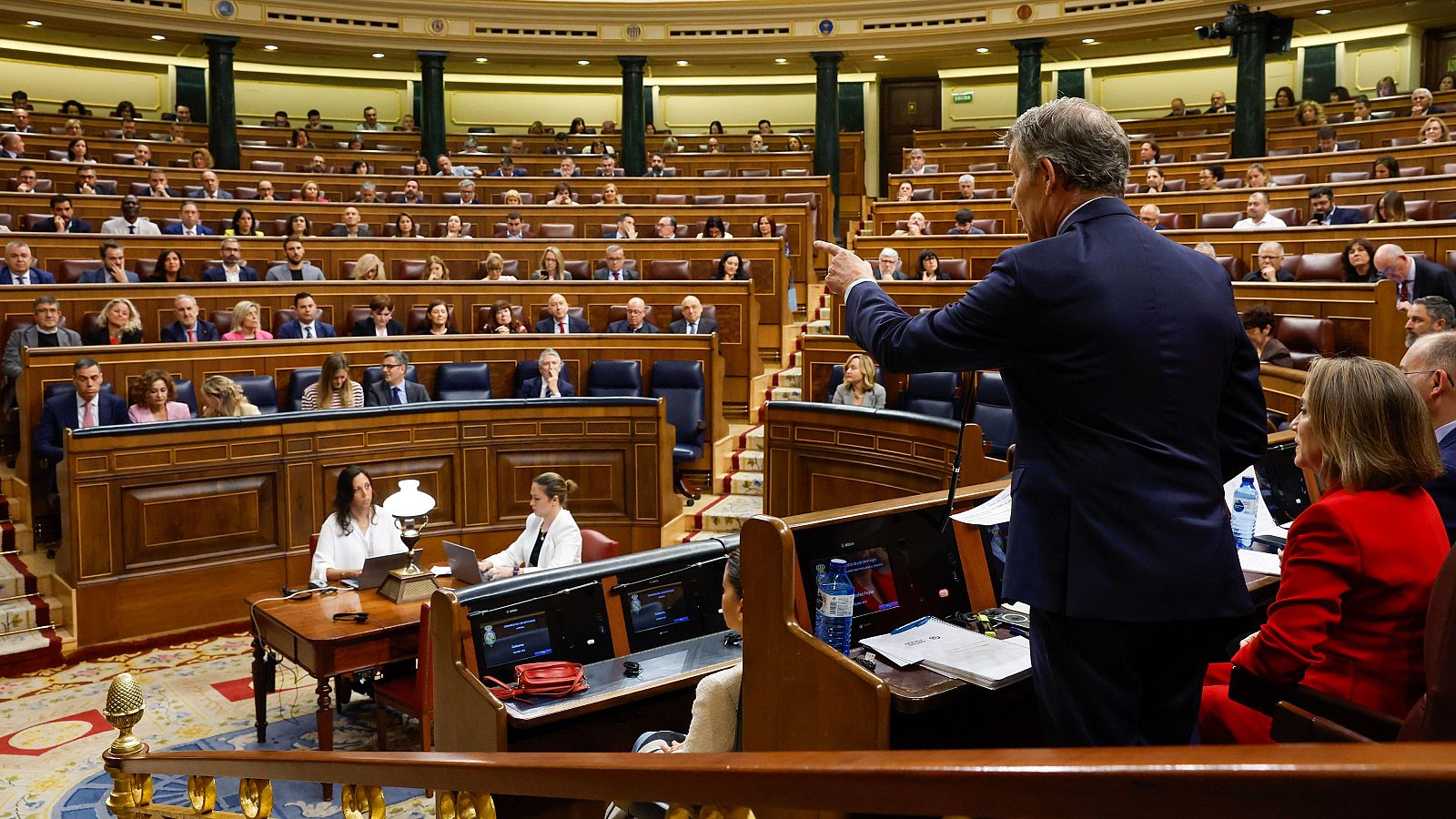 Sesión parlamentaria en el hemiciclo: miembro del gobierno gesticula desde la mesa, rodeado de diputados, diputadas y funcionarias trabajando con ordenadores.