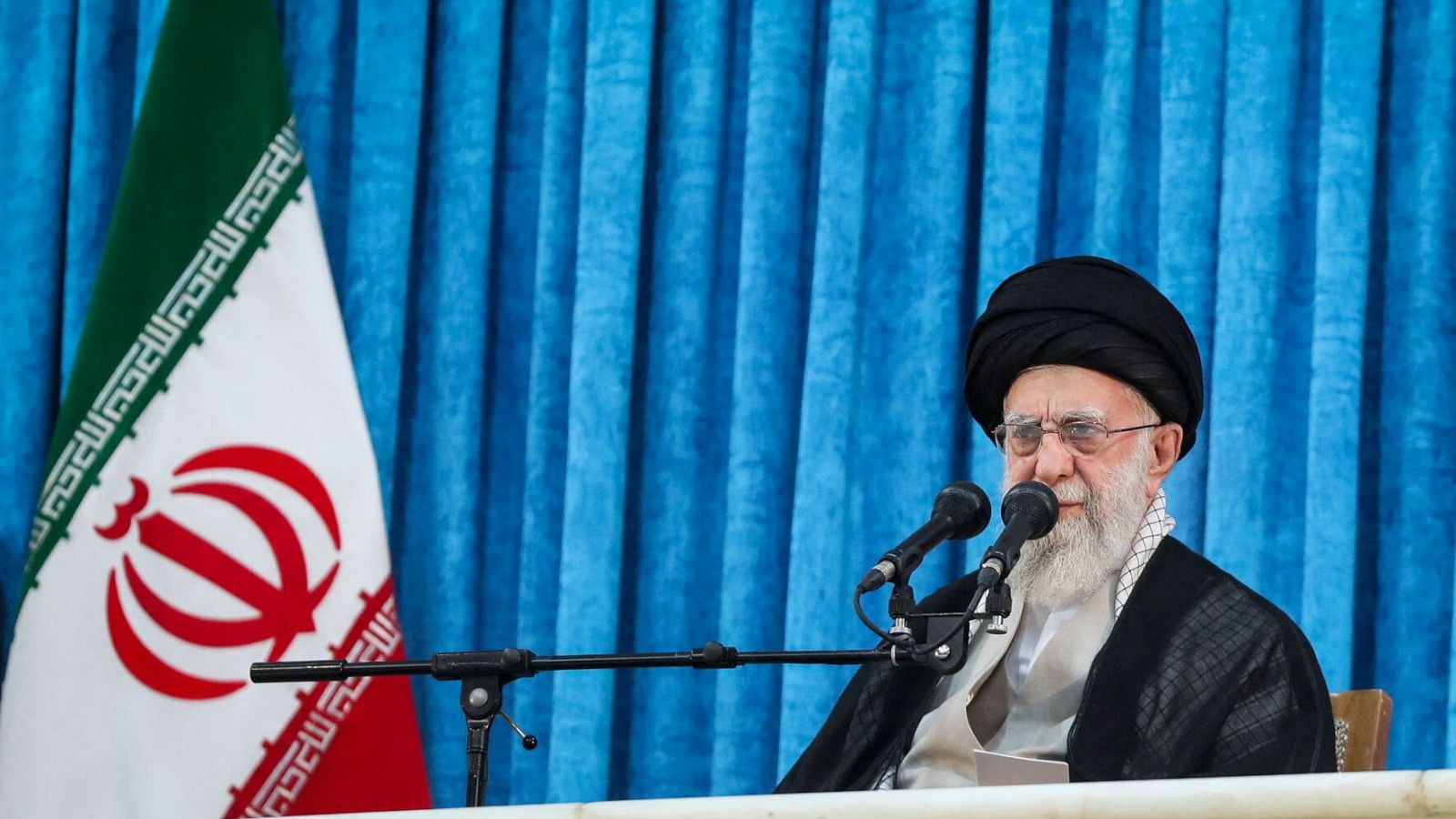 El Ayatolá Ali Jamenei, sentado y con turbante negro, ofrece un discurso ante dos micrófonos, con un telón azul y la bandera iraní al fondo.