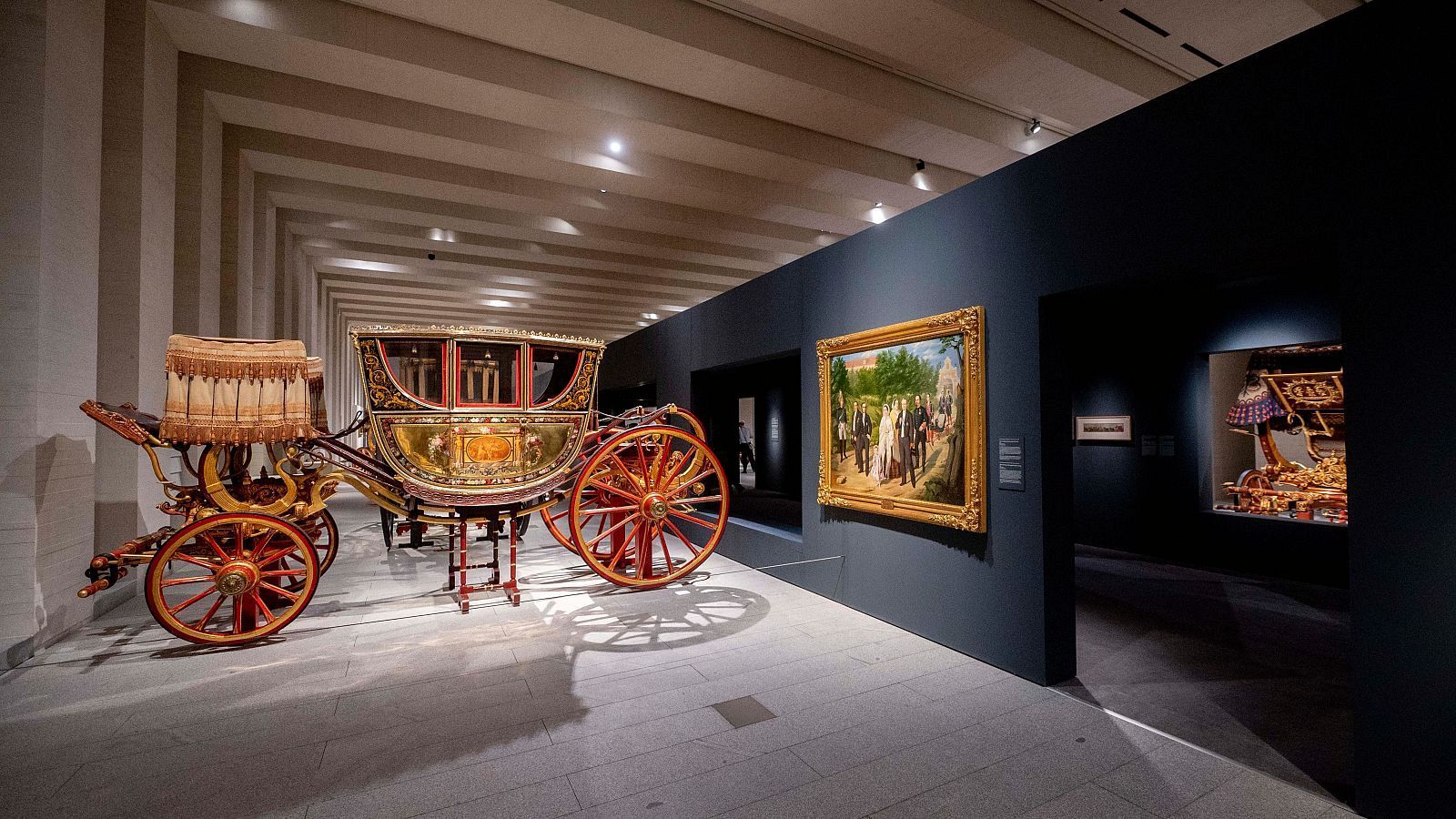 Imponente carroza roja y dorada con detalles ornamentales y ruedas de madera, expuesta en una sala amplia con iluminación focalizada. Se aprecia parte de otra pieza similar en una sala adyacente.