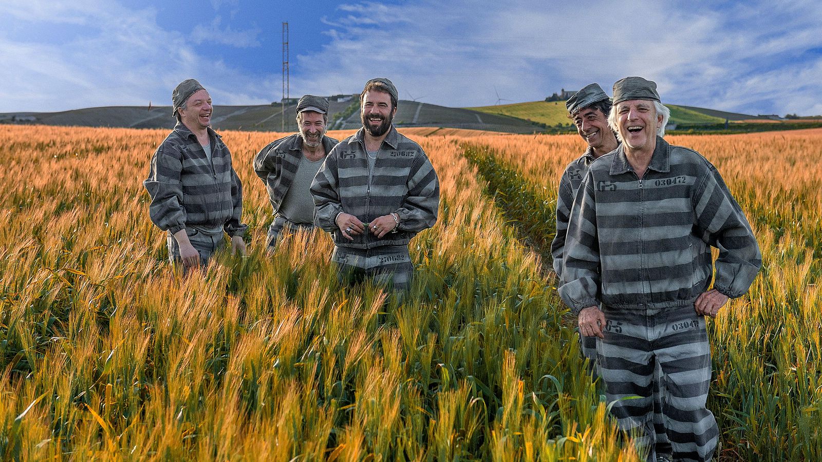 Cinco hombres, vestidos con uniformes de prisión grises, sonríen en un campo de trigo con colinas al fondo.  Imagen relacionada con la presentación de un nuevo disco musical.