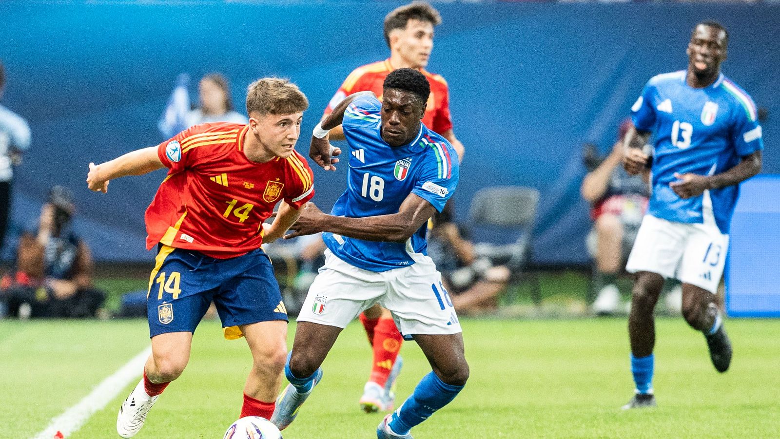 Partido de fútbol: un jugador español (14) con el balón, presionado por un jugador italiano (18).  Acción en el centro del campo.