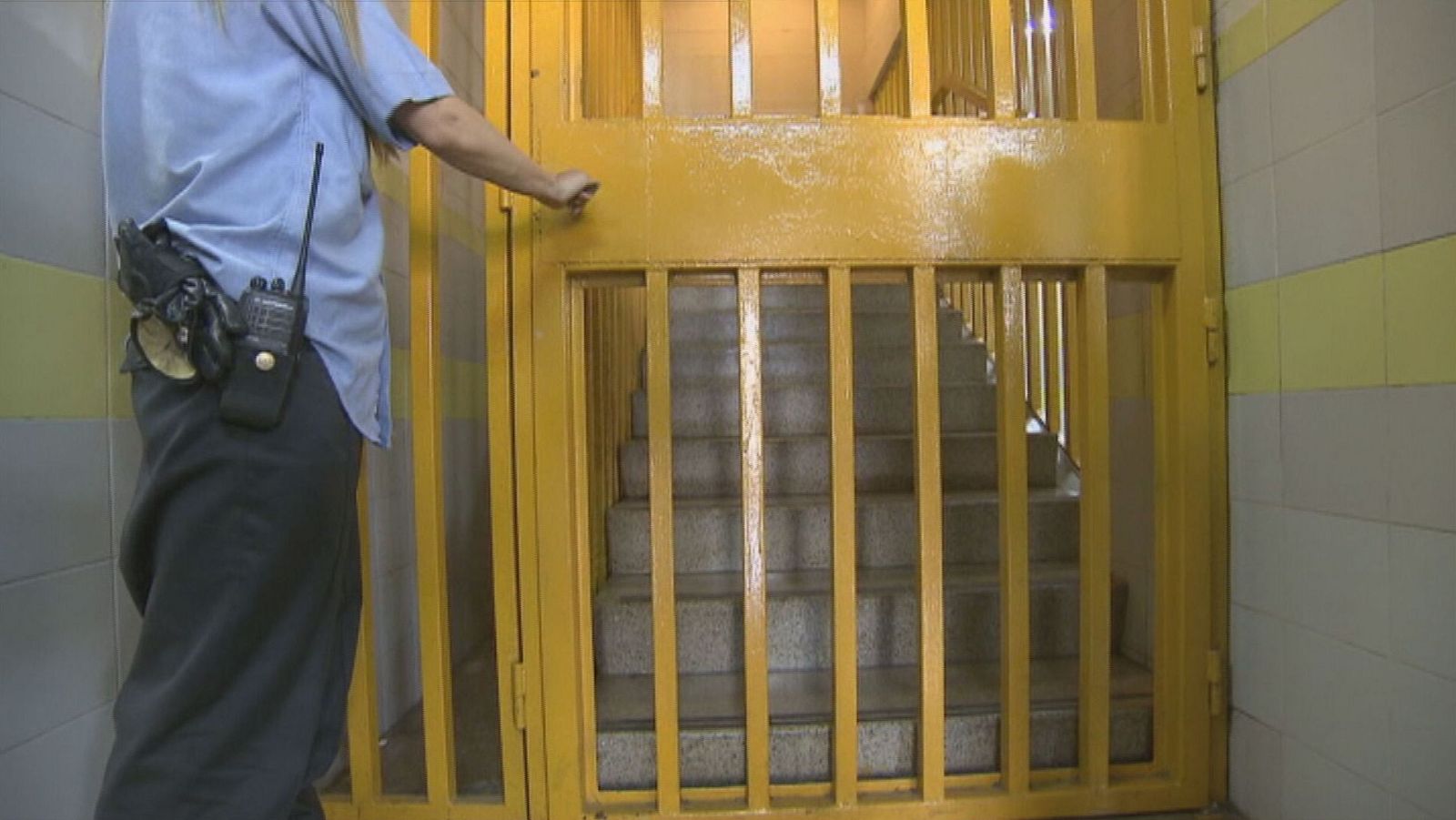 Funcionario de prisiones abre puerta con barrotes amarillos en pasillo con azulejos grises y amarillos, usando walkie-talkie y guantes.