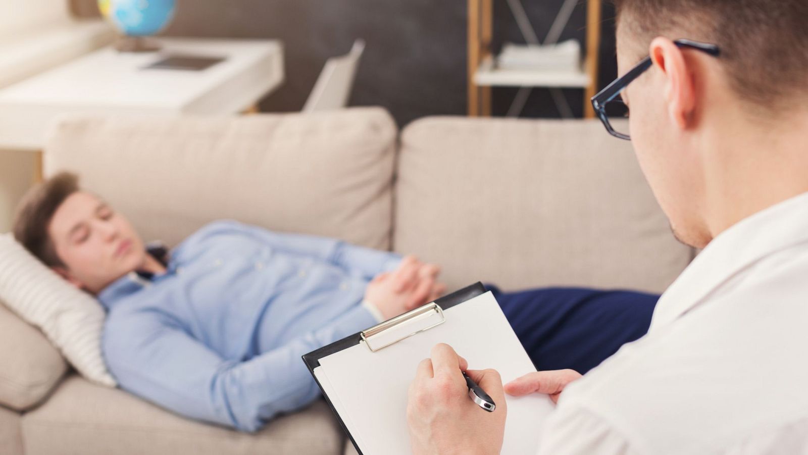 Joven en trance hipnótico en sofá, observado por terapeuta que toma notas en una consulta. Ambiente relajado.