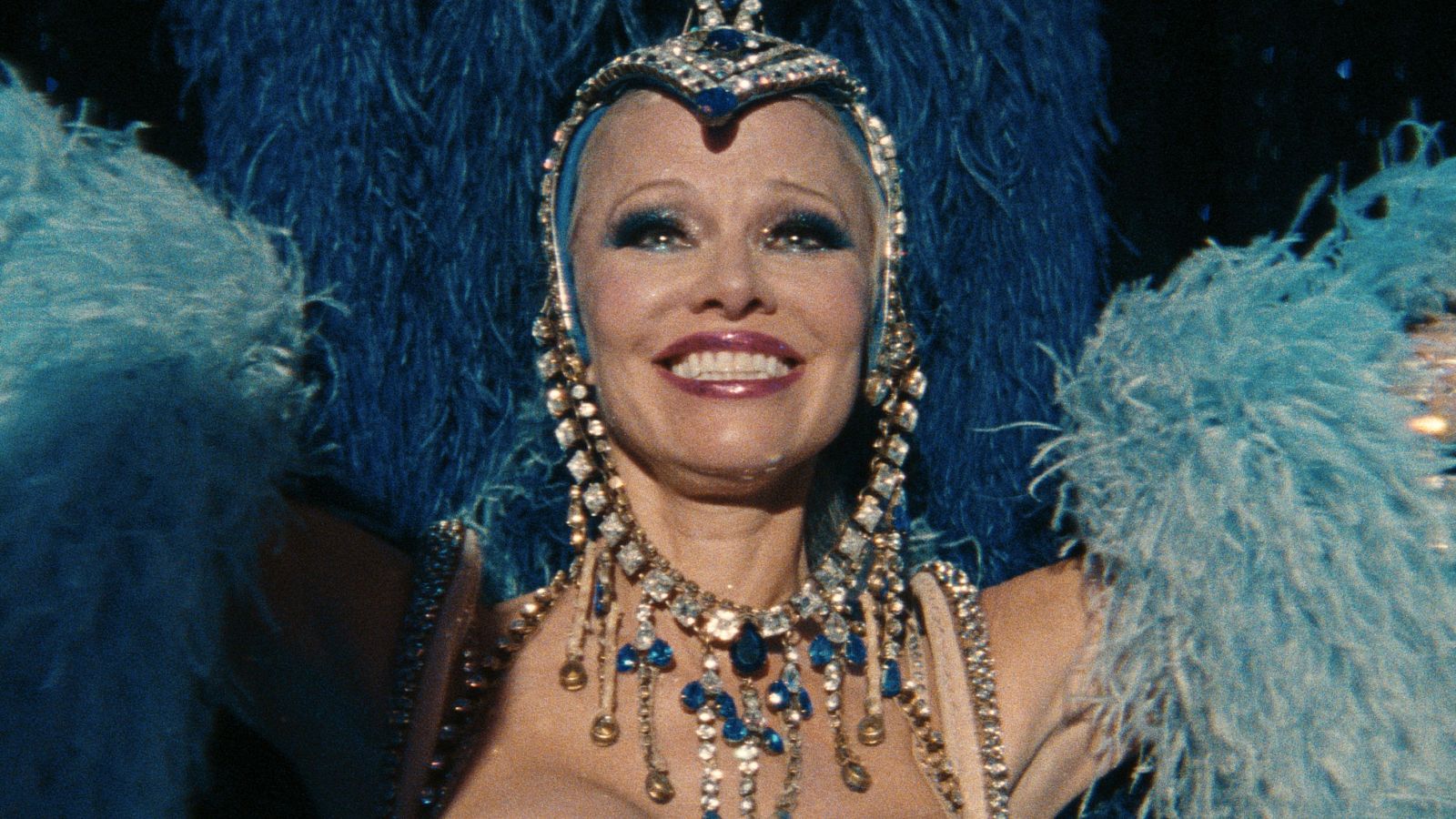 Imagen de Pamela Anderson con tocado azul, maquillaje intenso y collar con piedras azules, sonriendo en lo que parece una escena de 'The Last Showgirl'.