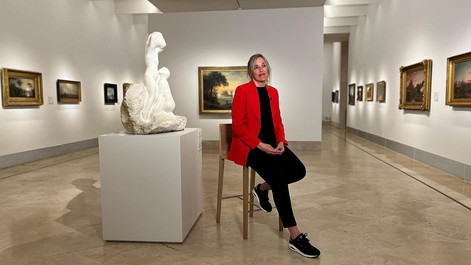Conservadora del museo sentada en taburete alto, con americana roja y pantalones negros, durante una entrevista.  Se aprecian cuadros en el fondo.