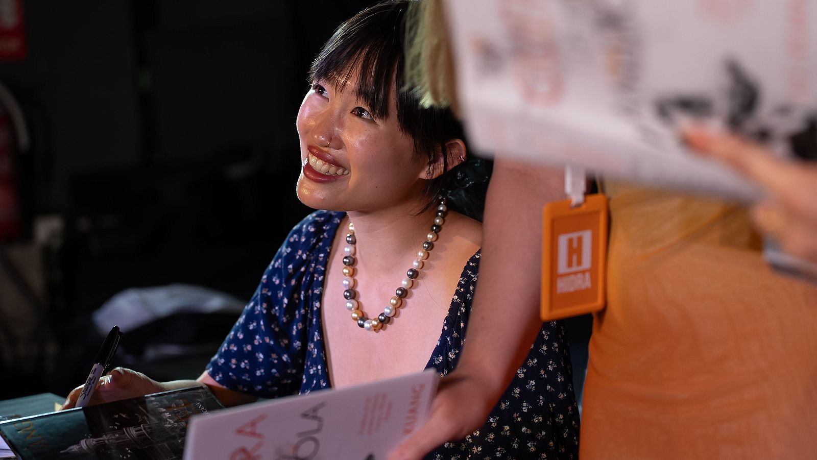 R. F. Kuang firma libros en la Feria del Libro de Madrid, sonriendo y vestida con un vestido azul oscuro con flores blancas y un collar de perlas. Se ve parte de la portada del libro y a otra persona con una acreditación naranja.