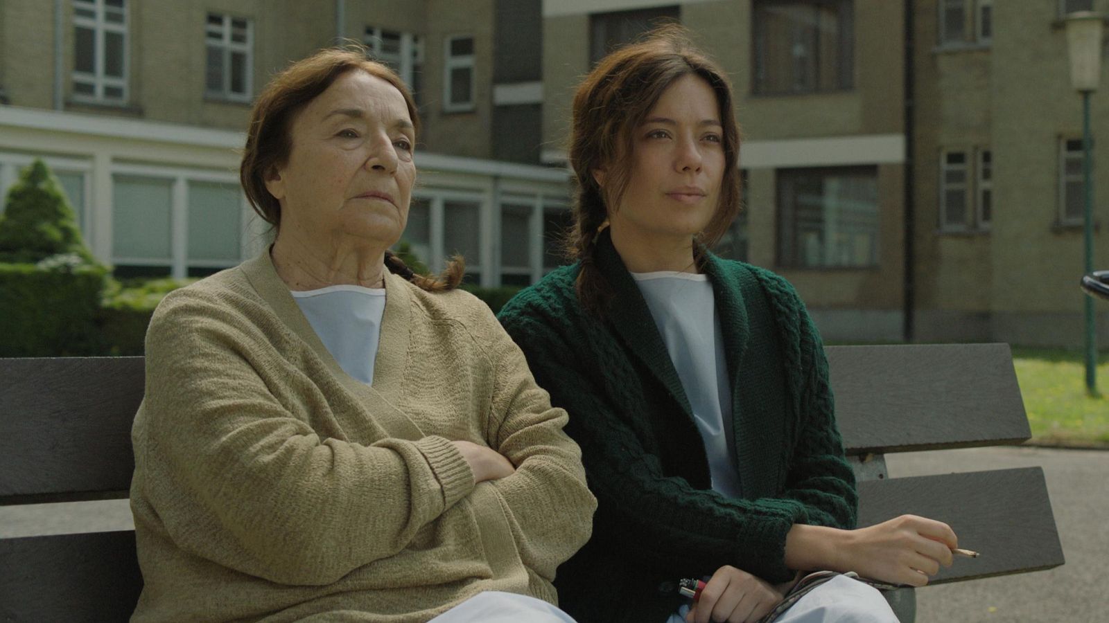 Dos mujeres, una mayor y otra joven con batas, sentadas en un banco exterior observan algo fuera de la imagen.  La escena parece un fotograma de una película.