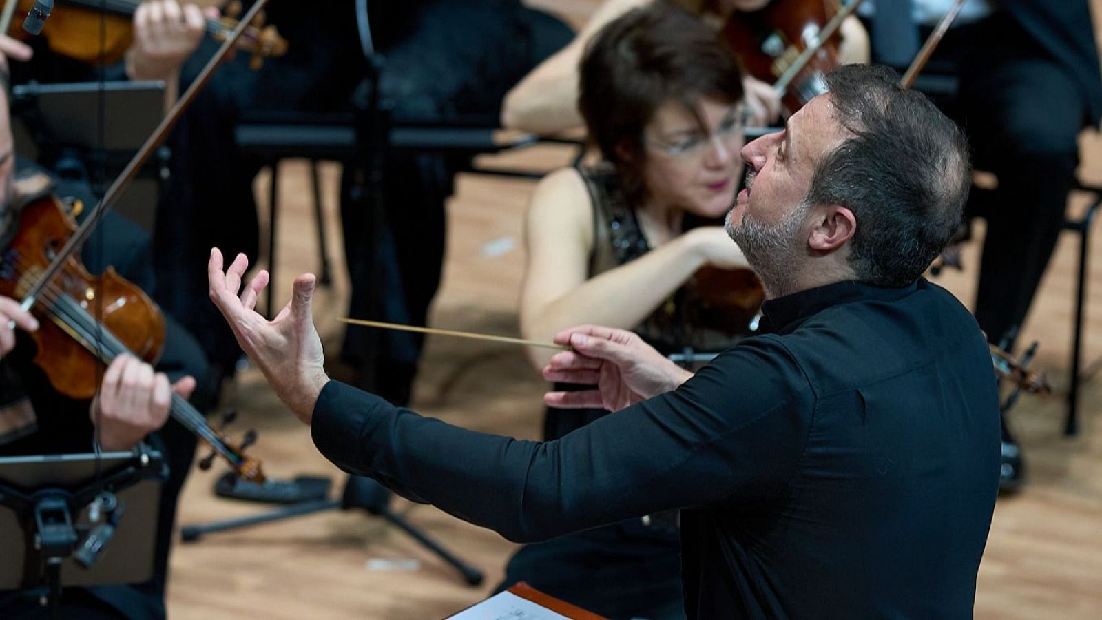 Director de orquesta en primer plano, con batuta en mano derecha y gestualizando con la izquierda, dirigiendo una orquesta con instrumentos de cuerda.