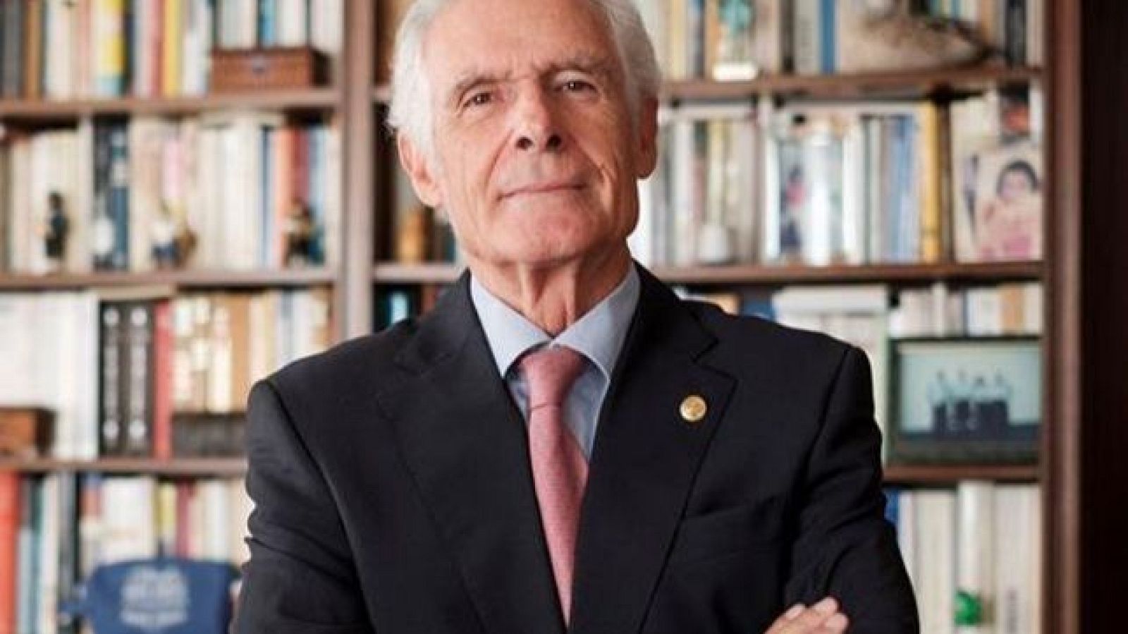 José María Aznar, en traje oscuro y corbata rosa, posa con los brazos cruzados frente a una estantería repleta de libros.  Pin en la solapa.