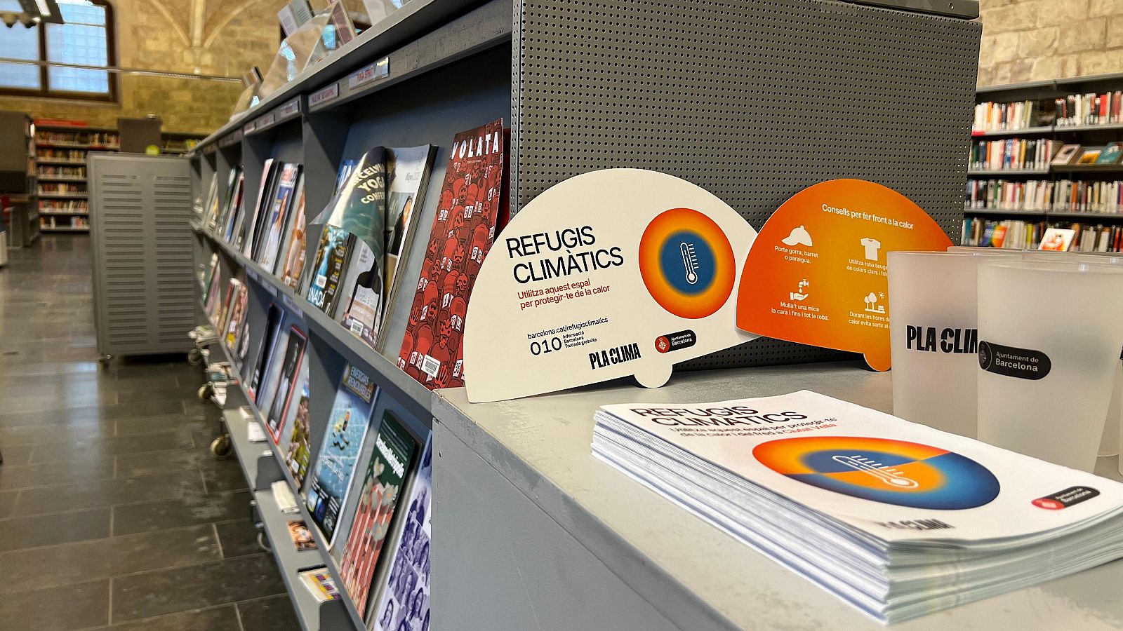 Material informativo sobre refugios climáticos en un mostrador de biblioteca: consejos para combatir el calor (ropa fresca, agua, etc.), logo "PLA CLIMA" y número de teléfono 010.