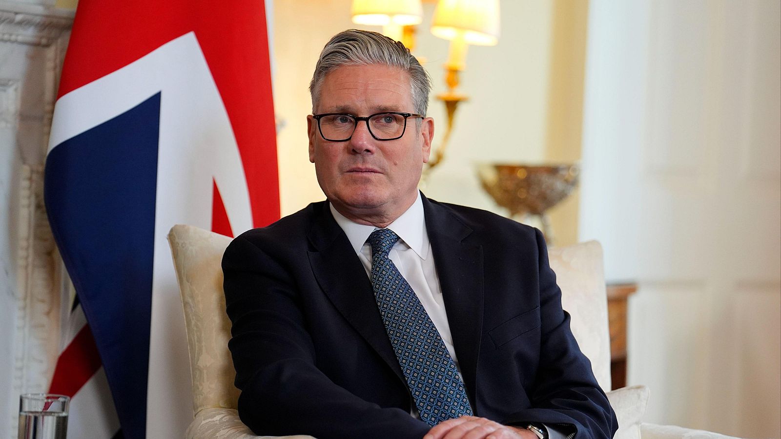 Fotografía de Keir Starmer, líder laborista británico, sentado en un sillón con traje oscuro y corbata geométrica; al fondo, una bandera del Reino Unido.
