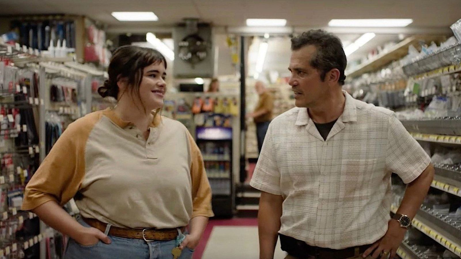 Escena en una ferretería: Benicio del Toro conversa con una mujer joven con camisa beige y pantalones vaqueros. Fondo con estanterías y gente.