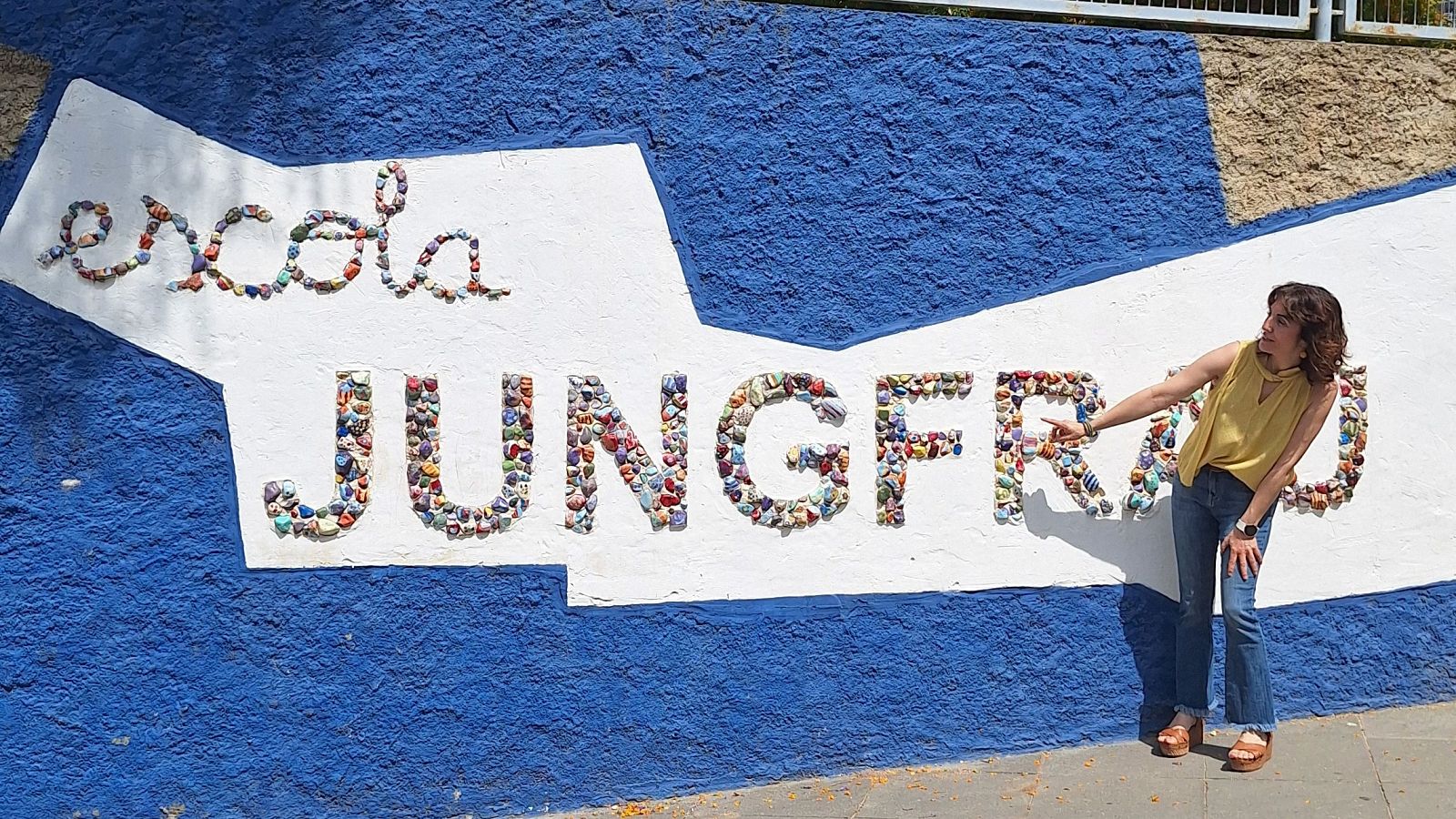 Mari Cruz Hernández a l'Escola Jungfrau de Badalona, participant en un programa especial de 'Lletra Lligada' de Ràdio 4 sobre educació juvenil. Senyala un cartell mosaic de colors.