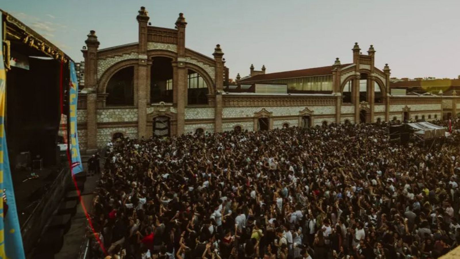 Concierto multitudinario al atardecer en Matadero Madrid.  El escenario está decorado con banderas y la gente disfruta del evento con los brazos levantados.
