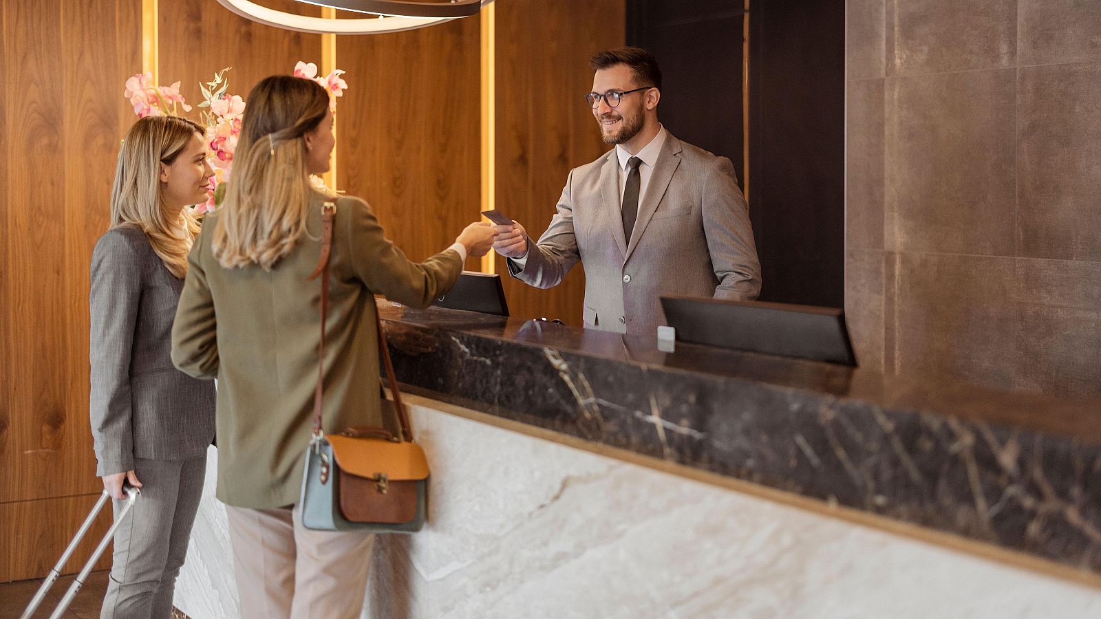 Recepción de hotel: recepcionista entrega tarjeta a mujer con camisa verde oliva y pantalones beige; otra mujer espera con traje gris y maleta.