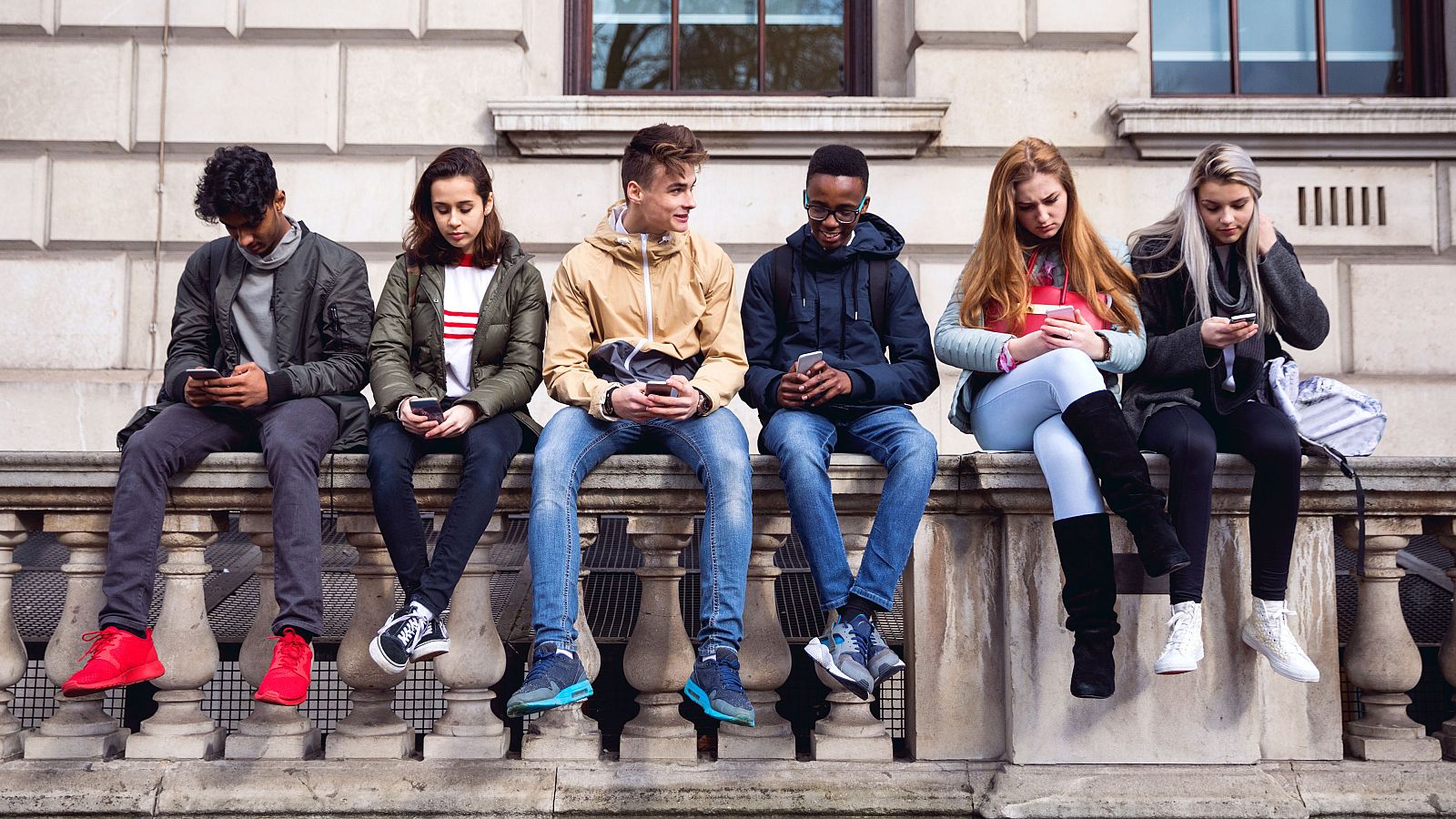 Seis adolescentes, tres chicos y tres chicas con ropa informal, miran sus móviles sentados en una barandilla de un espacio público.