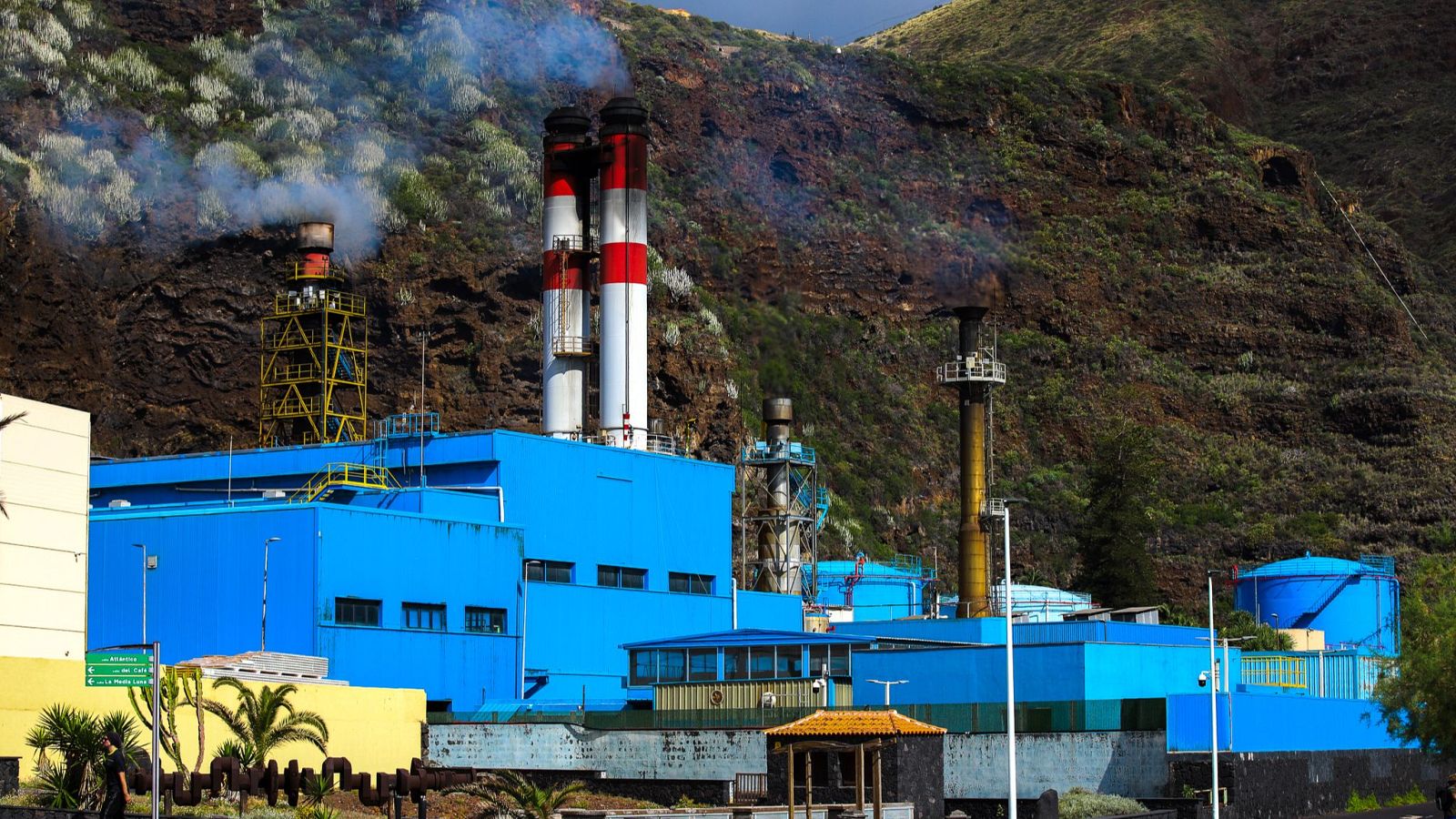 Central eléctrica en funcionamiento, al pie de una montaña con vegetación.  Se observa humo en algunas chimeneas y una zona de descanso con palmeras en primer plano.