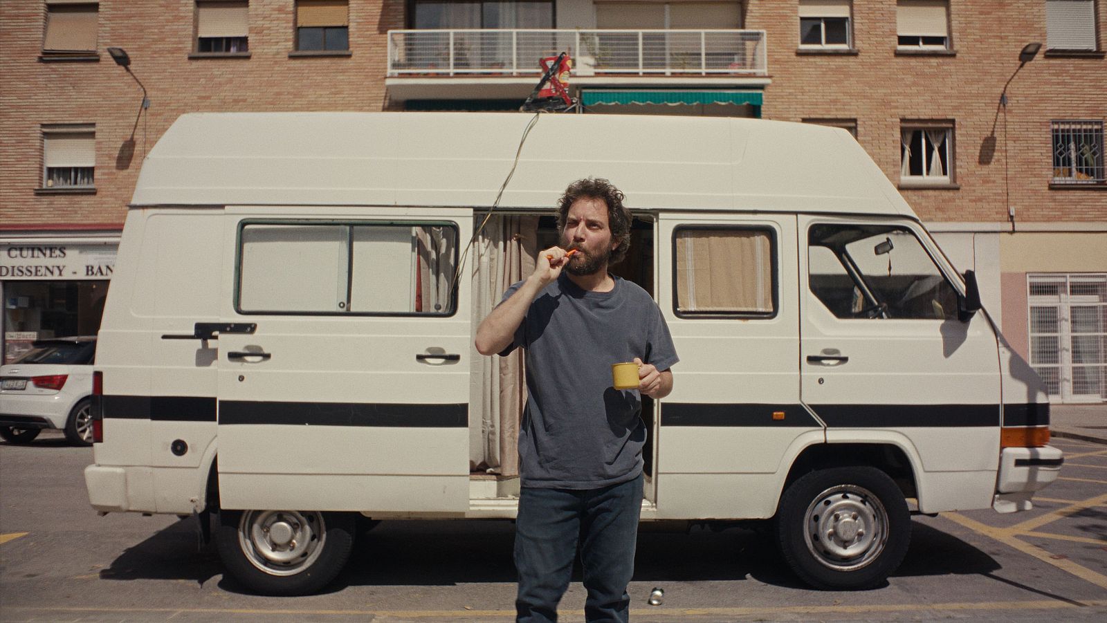 Un hombre con barba y pelo rizado cepilla sus dientes junto a una furgoneta blanca aparcada;  la puerta lateral está abierta y se ve una antena en el techo.