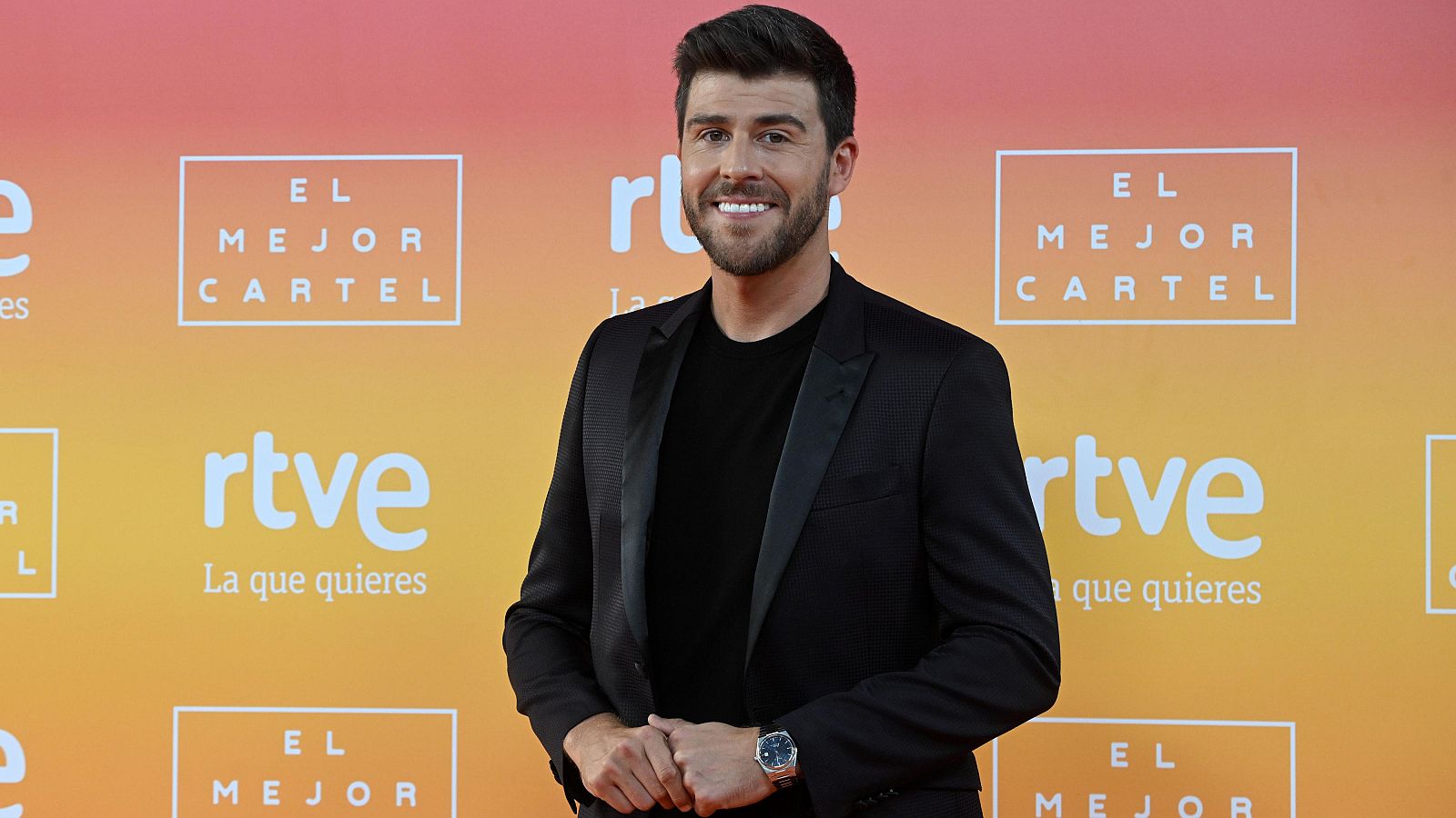 Presentador de RTVE en presentación de nueva programación. Chaqueta negra, fondo naranja y amarillo con logo RTVE y el eslogan "La que quieres".