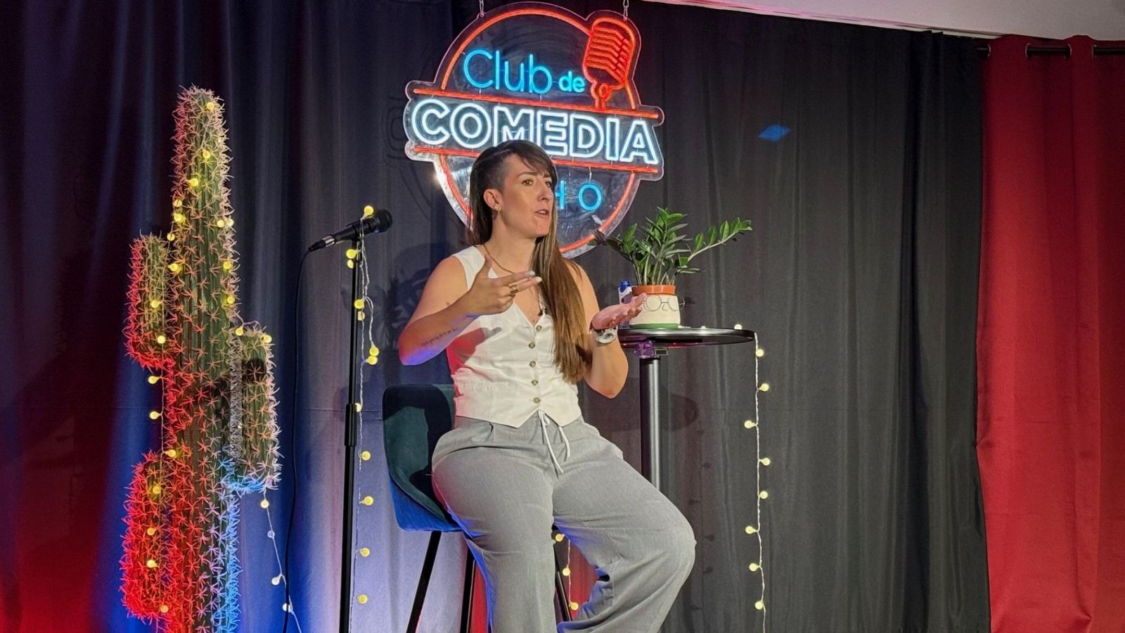 Comediante en entrevista, gesticulando con las manos frente a un micrófono en un escenario con plantas y luces de neón.