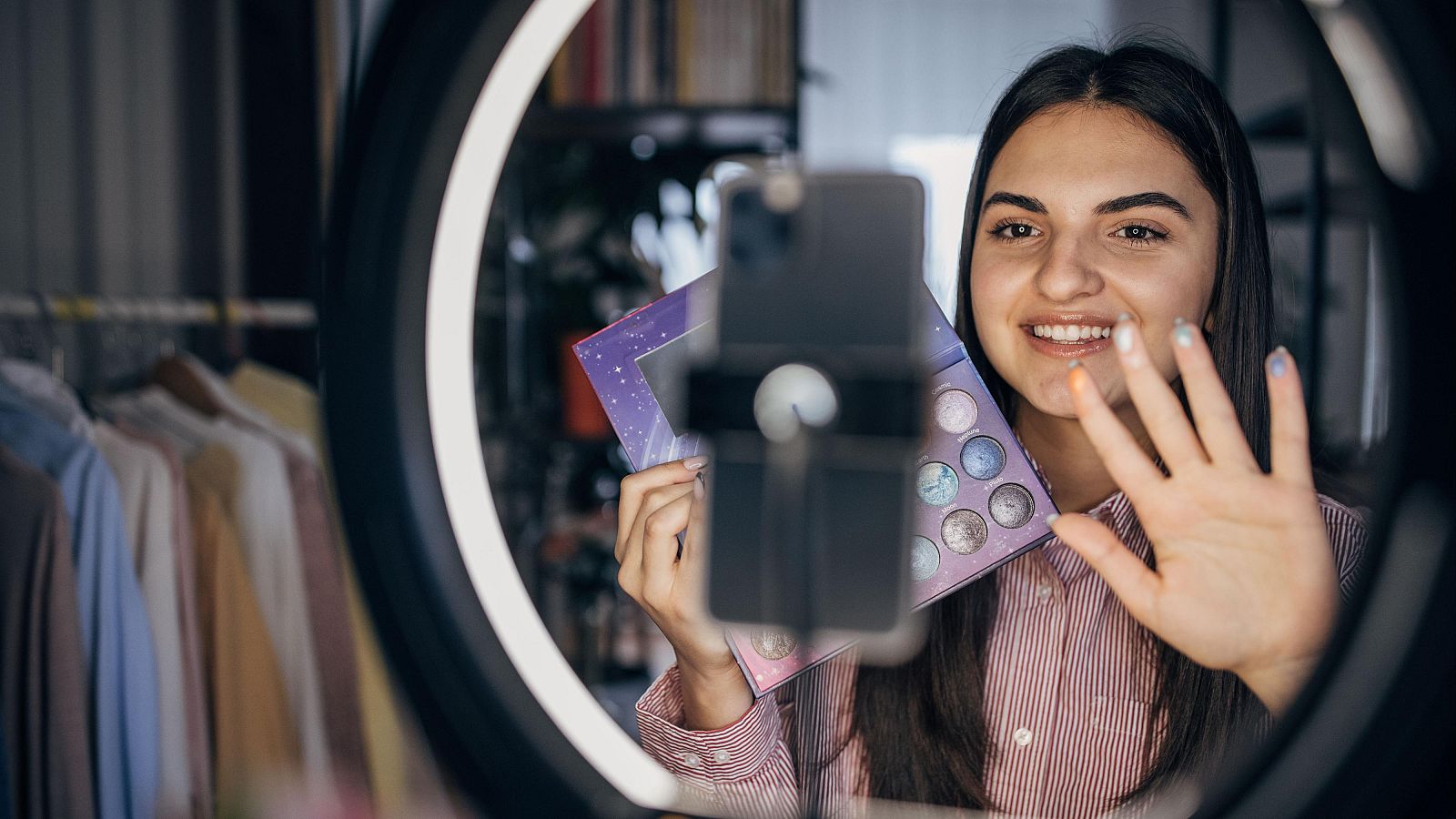 Mujer sonriente promociona una paleta de sombras de ojos en un vídeo, usando un aro de luz y vestida con una camisa a rayas. Fondo con ropa colgada.