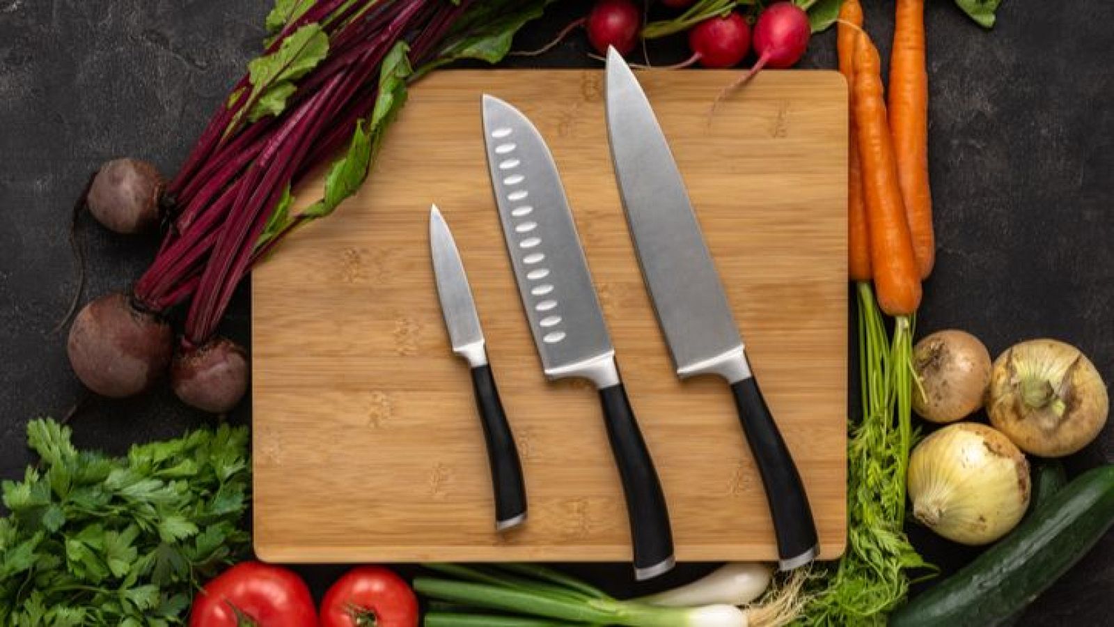 Tres cuchillos (chef, mediano y pelar) sobre tabla de madera, rodeados de verduras frescas: remolachas, zanahorias, cebollas, pepinos, tomates y perejil.