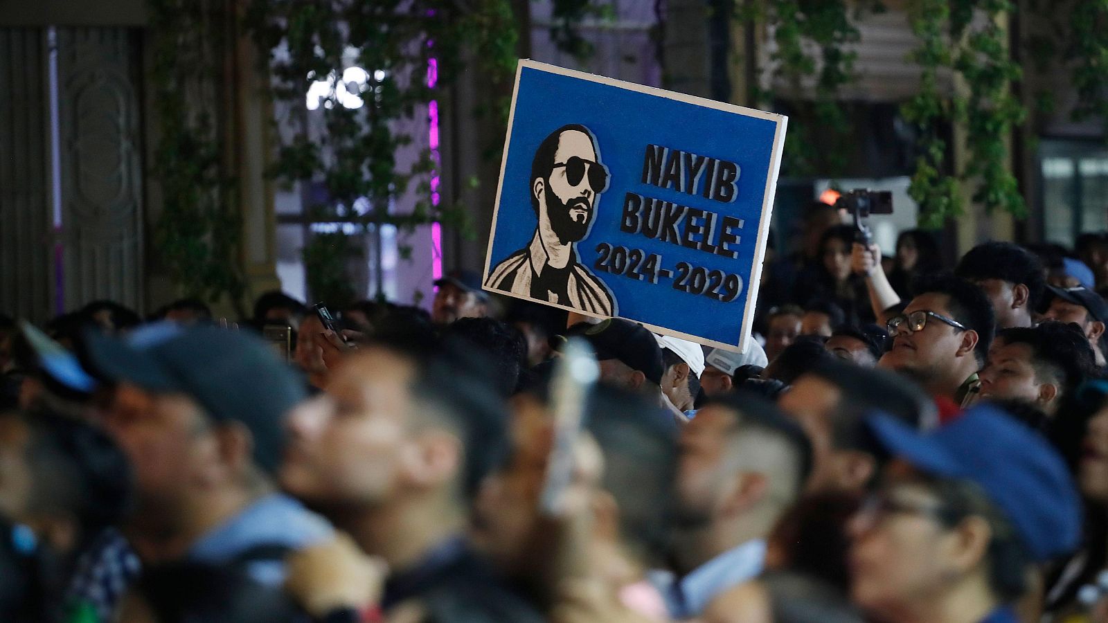 Cartel azul con imagen de Nayib Bukele y el texto "NAYIB BUKELE: 2024-2029", sostenido entre una multitud en un evento público.