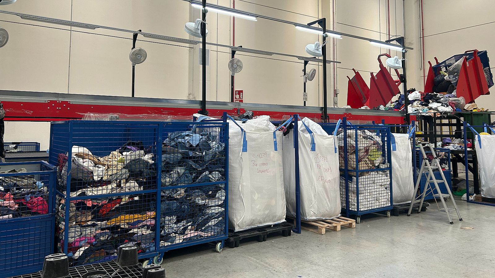 Cinta transportadora roja moviendo ropa a contenedores azules y sacos blancos en una gran nave industrial.  Posible centro de clasificación o almacenamiento textil.