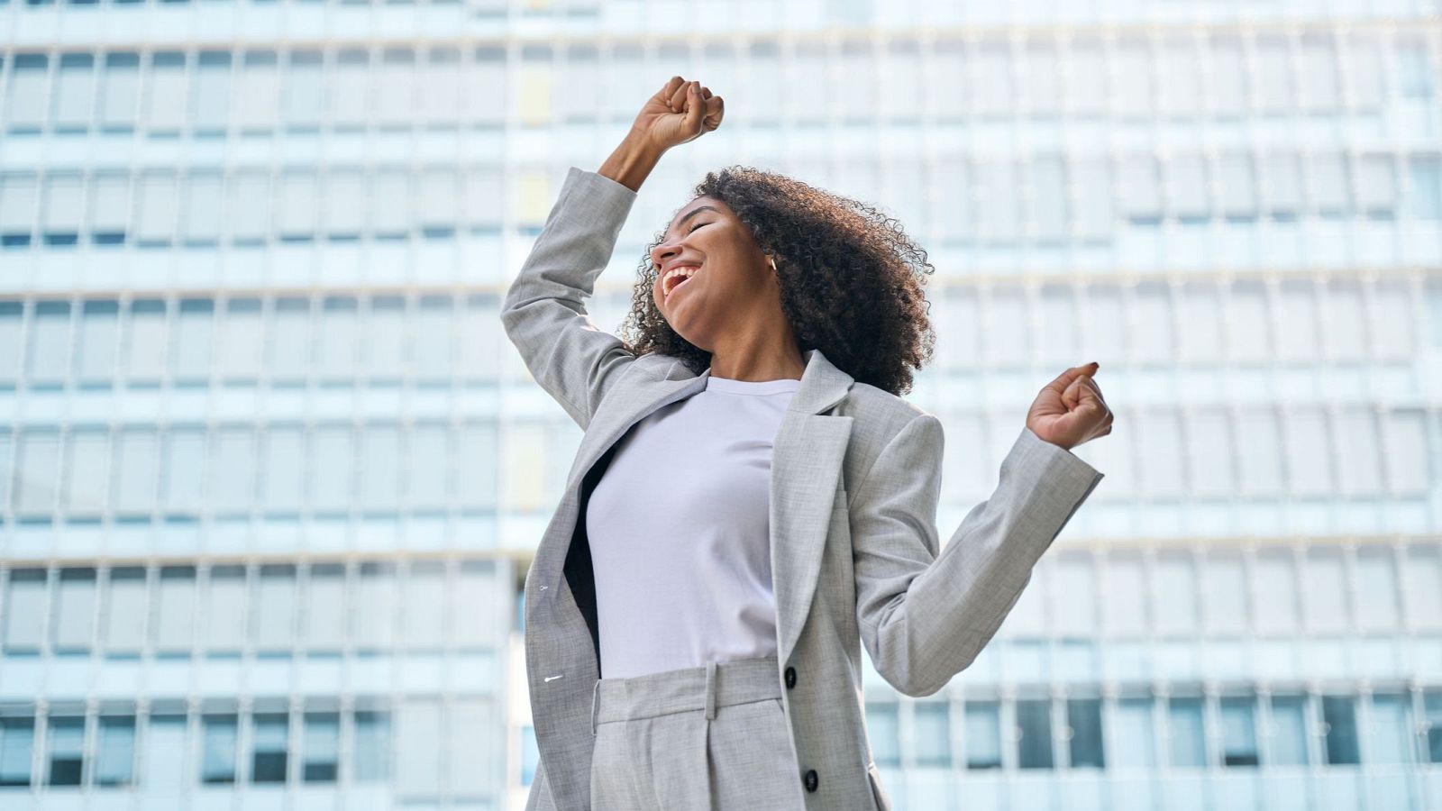 Mujer joven en traje gris celebra un logro con puños en alto, mostrando alegría en un edificio moderno de oficinas.