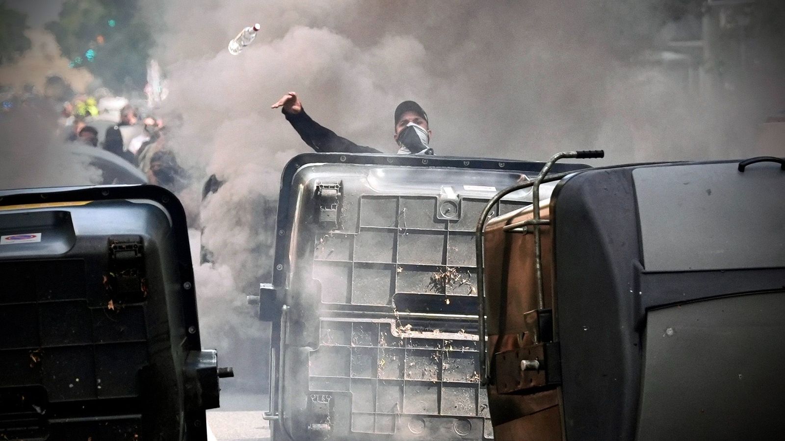 Hombre lanzando botella en una protesta con humo y contenedores volcados como barricadas.  Tensión y confrontación.
