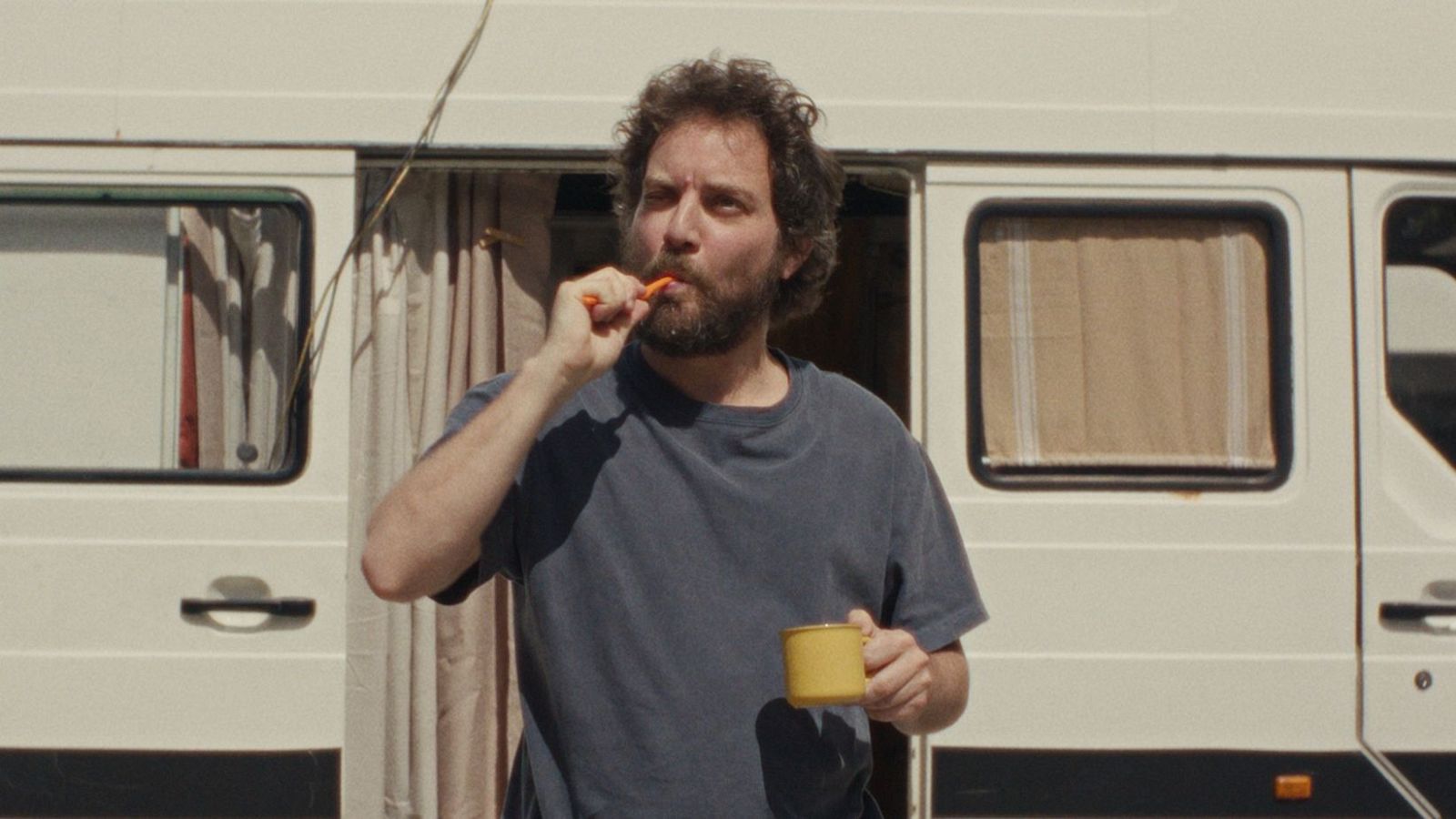 Actor con barba y pelo rizado cepillándose los dientes con un cepillo naranja, sosteniendo una taza amarilla, frente a una furgoneta blanca con la puerta abierta.