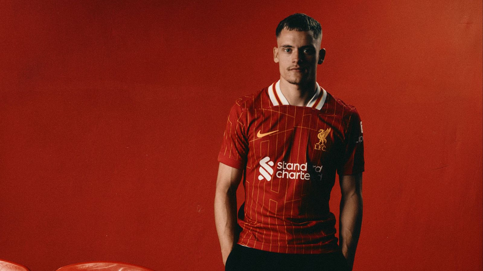 Florian Wirtz posa con la camiseta roja del Liverpool, mostrando los logos de Nike y Standard Chartered. Pelo corto y rubio, mirada directa a cámara.