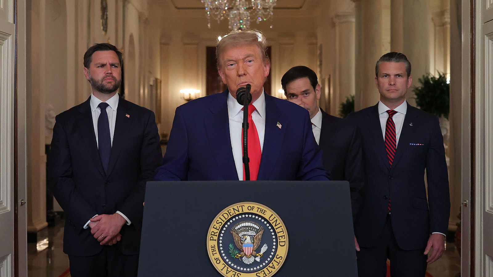 Discurso presidencial en la Casa Blanca. El expresidente Trump, en traje azul y corbata roja, habla desde un atril con el sello presidencial. Tres hombres en traje lo acompañan.