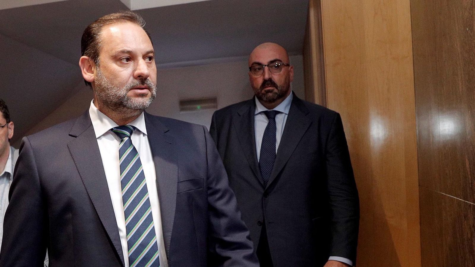 Fotografía de dos hombres en traje, uno en primer plano y otro ligeramente desenfocado al fondo, posiblemente en un pasillo o entrada.  Uno de ellos tiene barba y corbata a rayas.