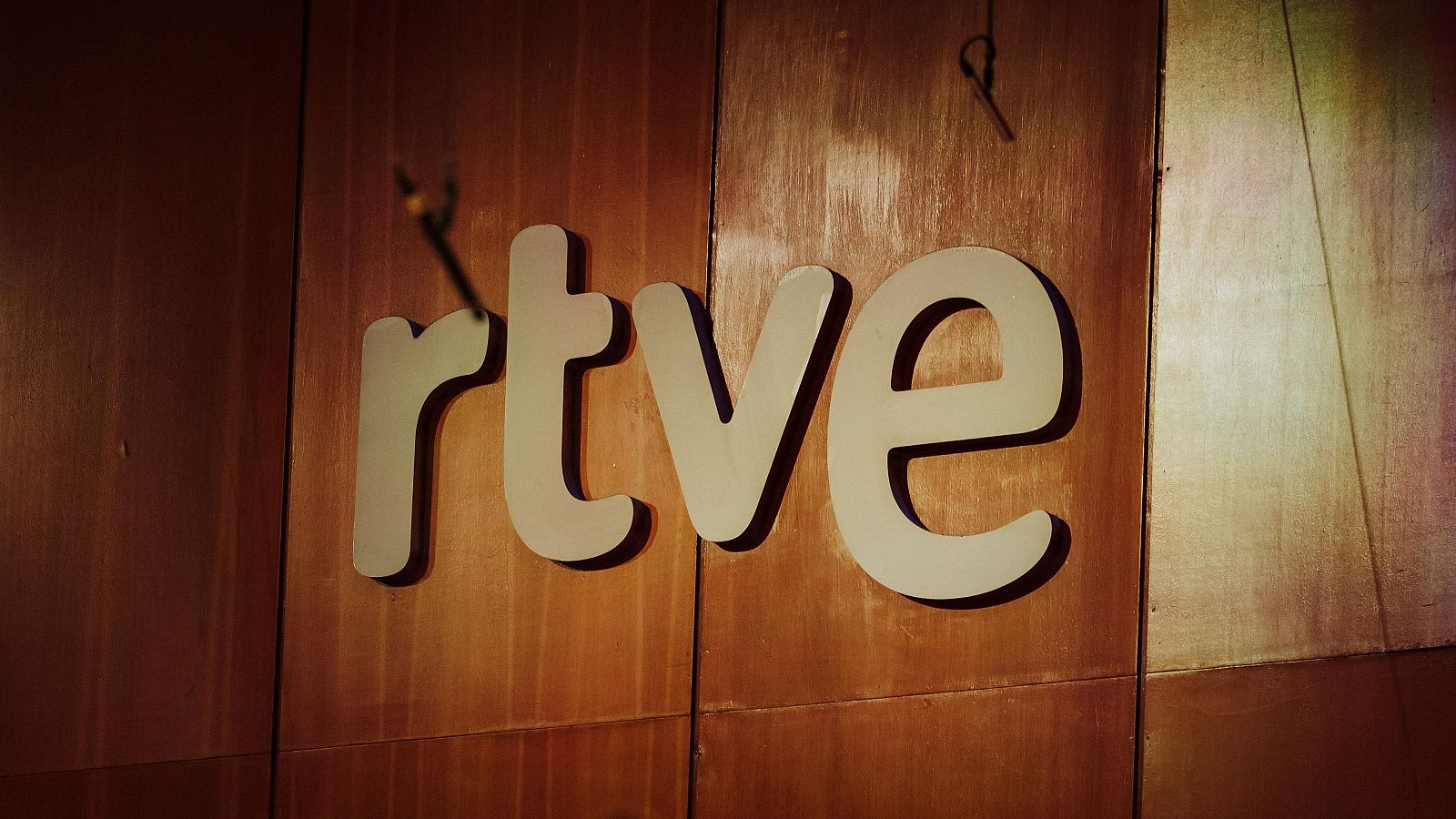Logotipo RTVE en relieve, color beige claro, sobre madera oscura con vetas.  Objetos oscuros, posiblemente cables, visibles arriba.