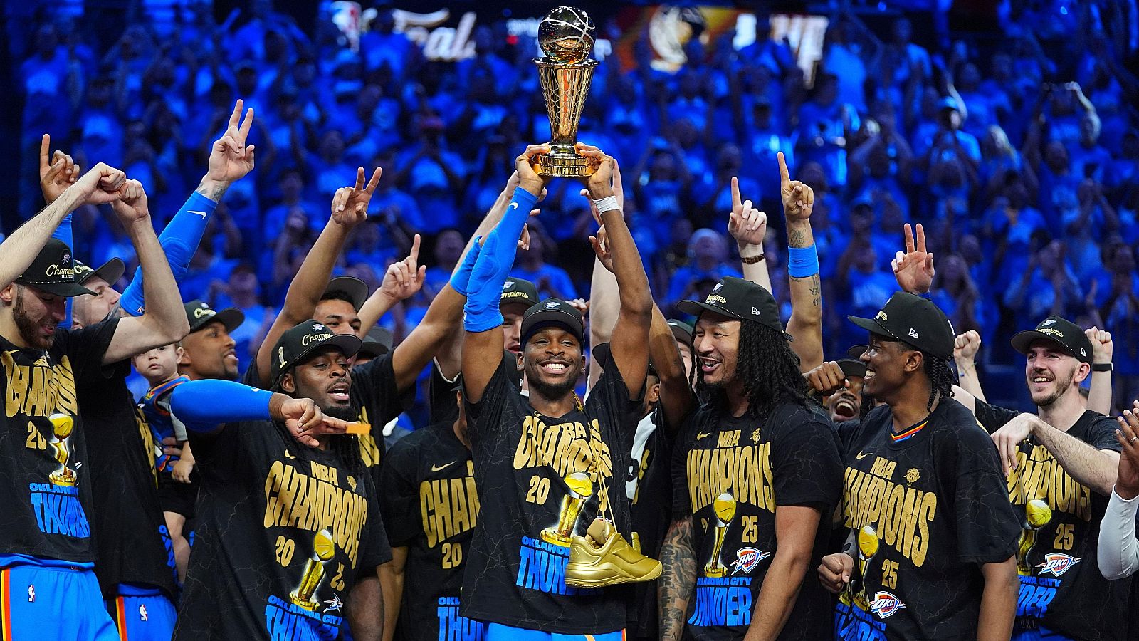 Jugadores de baloncesto celebran victoria, alzando trofeo. Visten camisetas y gorras negras con "NBA CHAMPIONS" en dorado.  Alegría y euforia.