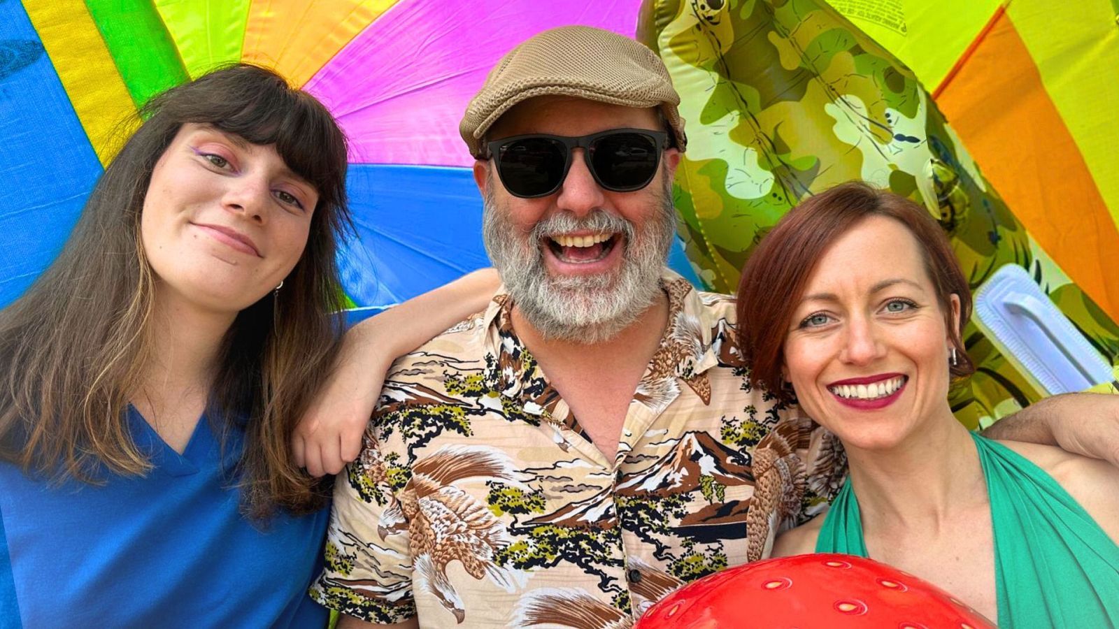 Tres personas posan para una foto promocional veraniega: una mujer con camiseta azul, un hombre con gorra y gafas, y una mujer pelirroja con vestido verde y pelota de playa. Ambiente festivo y soleado.