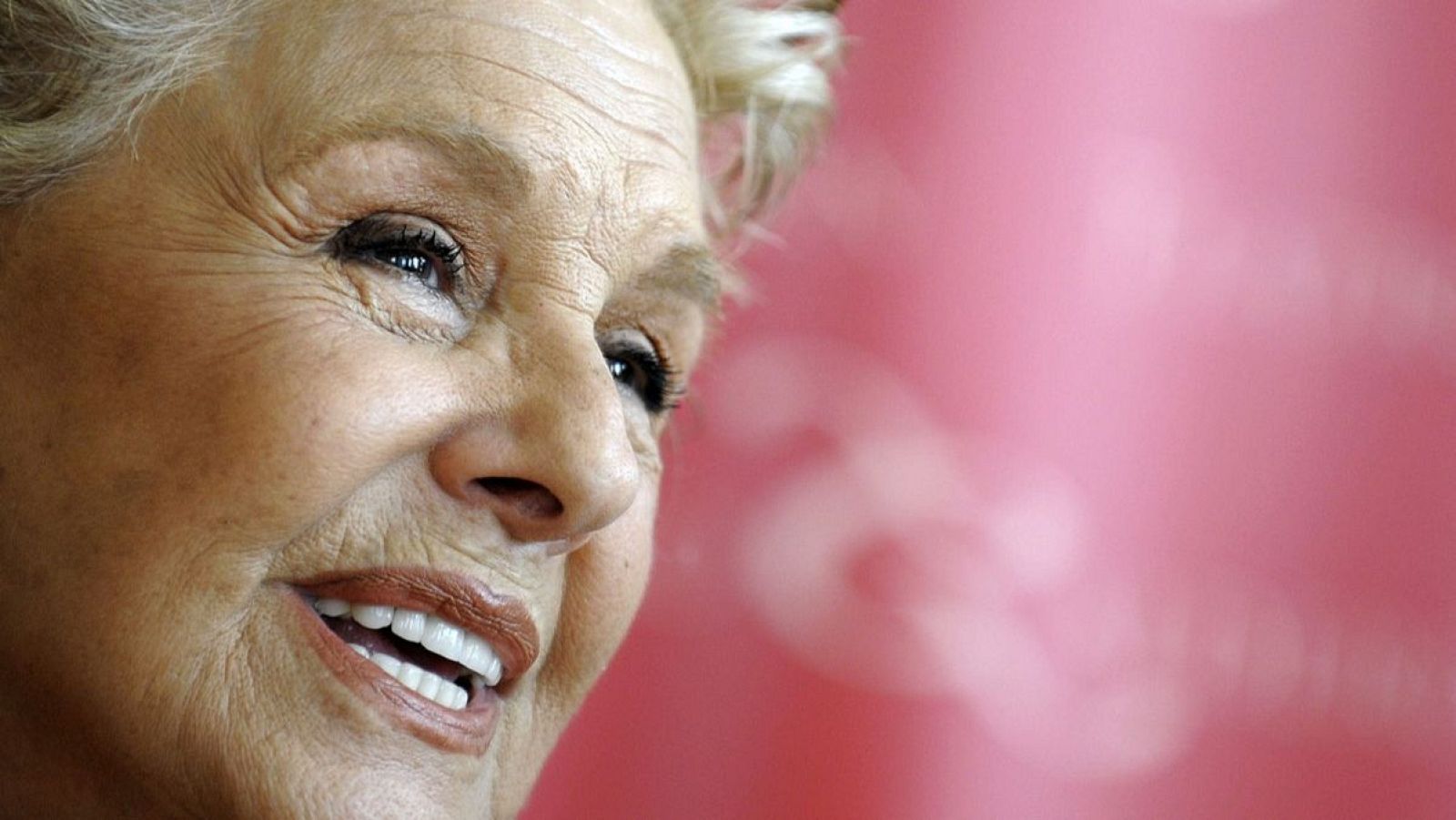 Primer plano de Concha Velasco sonriendo, con cabello rubio corto y canoso, mostrando arrugas. Fondo rosa claro desenfocado.