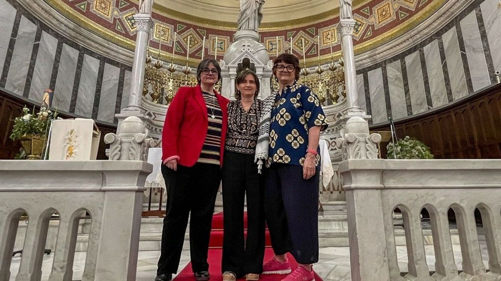 Foto de tres mujeres con atuendos informales posando en una iglesia sobre una alfombra roja tras unas entrevistas. Fondo con balaustrada de mármol y altar.