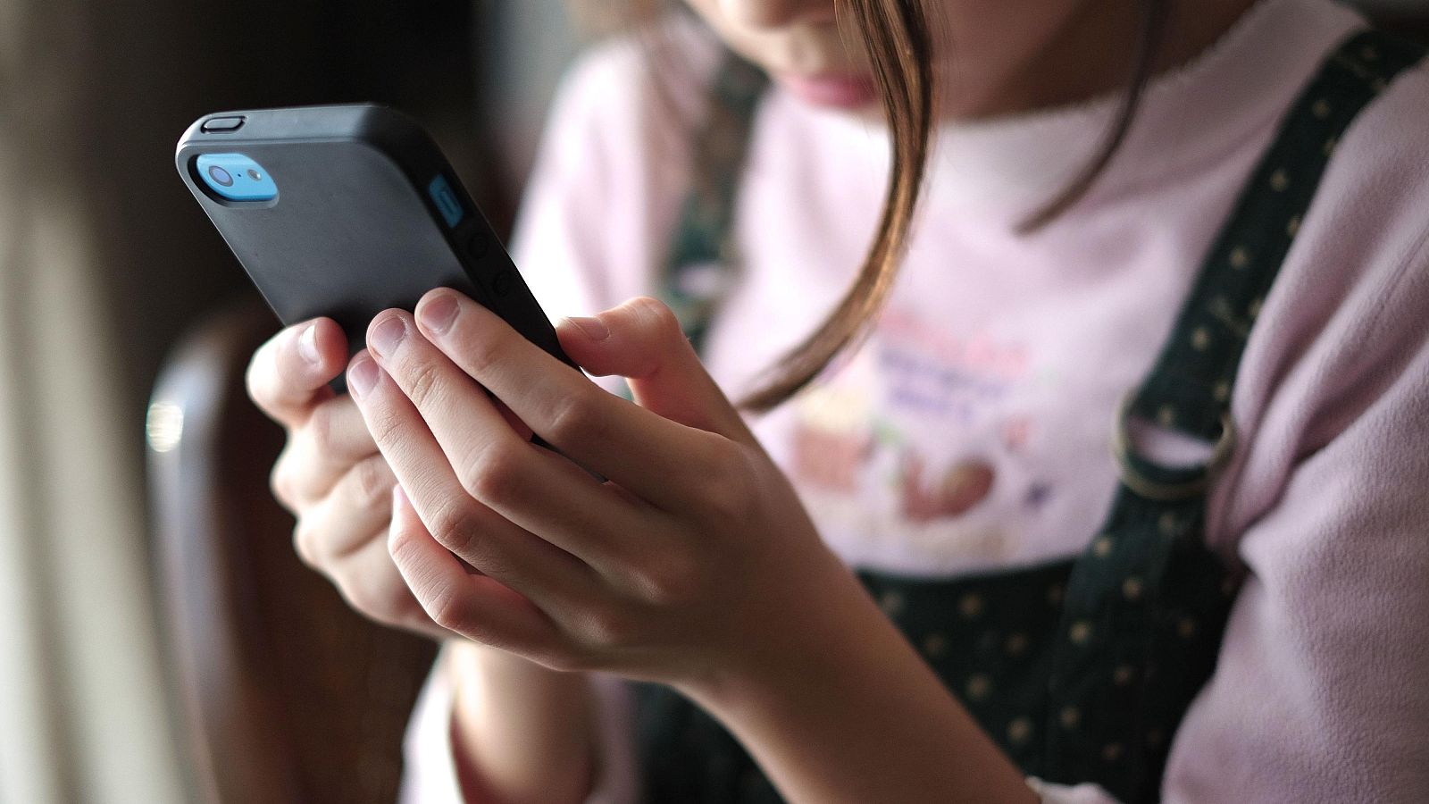 Niña con vestido rosa y móvil con funda negra, vista de cerca, manos interactuando con la pantalla, rostro desenfocado, iluminación interior.