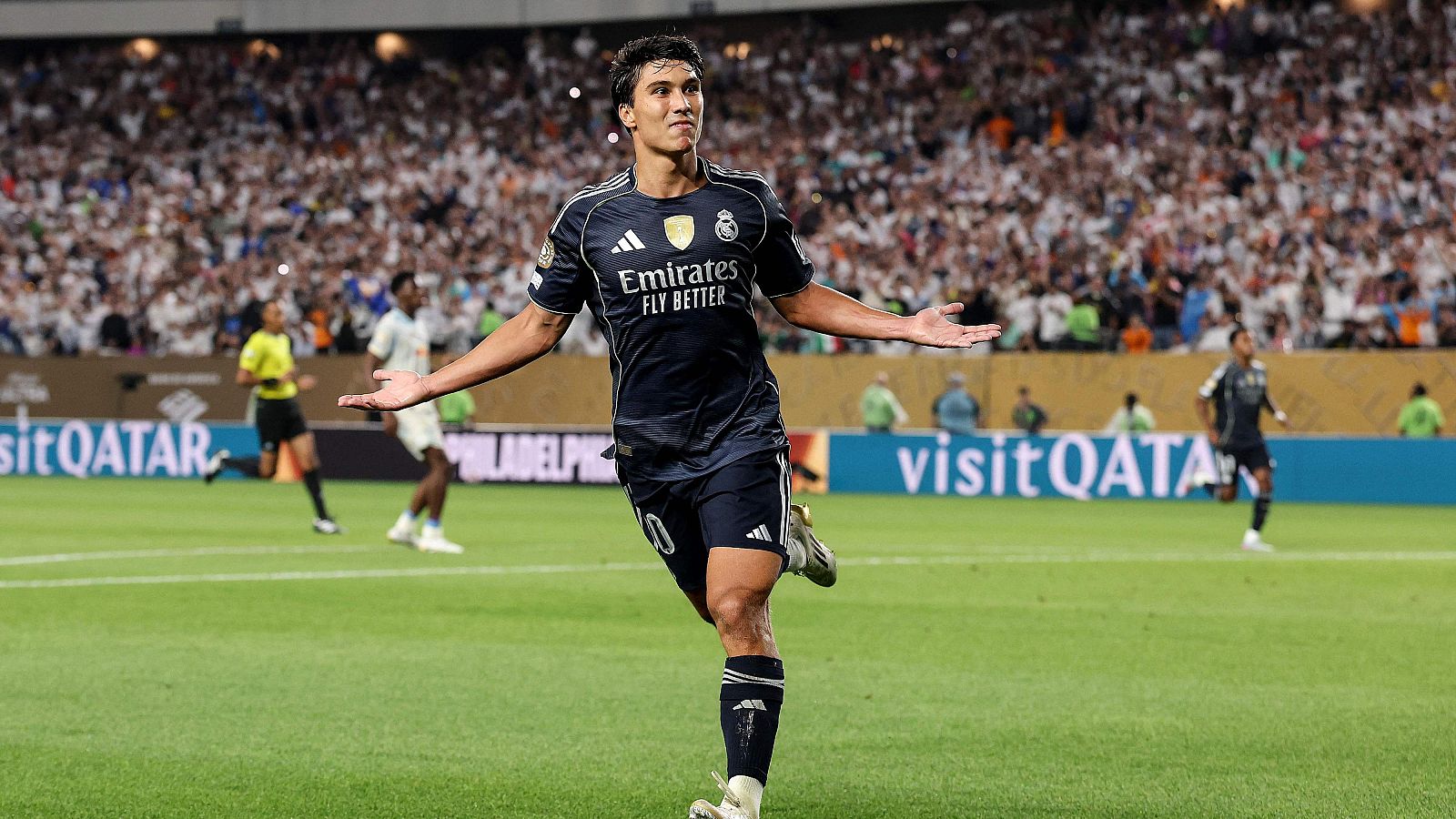 Celebración de gol de un jugador con camiseta negra del Real Madrid y patrocinador Emirates. Estadio lleno, césped en buen estado.