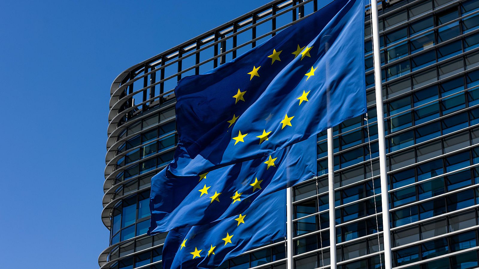 Banderas europeas ondeando ante un edificio de oficinas moderno con fachada de cristal y estructura de acero visible.