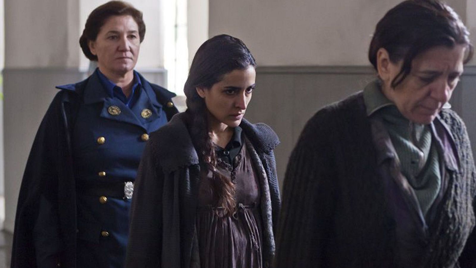 Tres mujeres con ropa de época, una de ellas con uniforme oscuro, en una escena dramática de interior.  Atmósfera seria.
