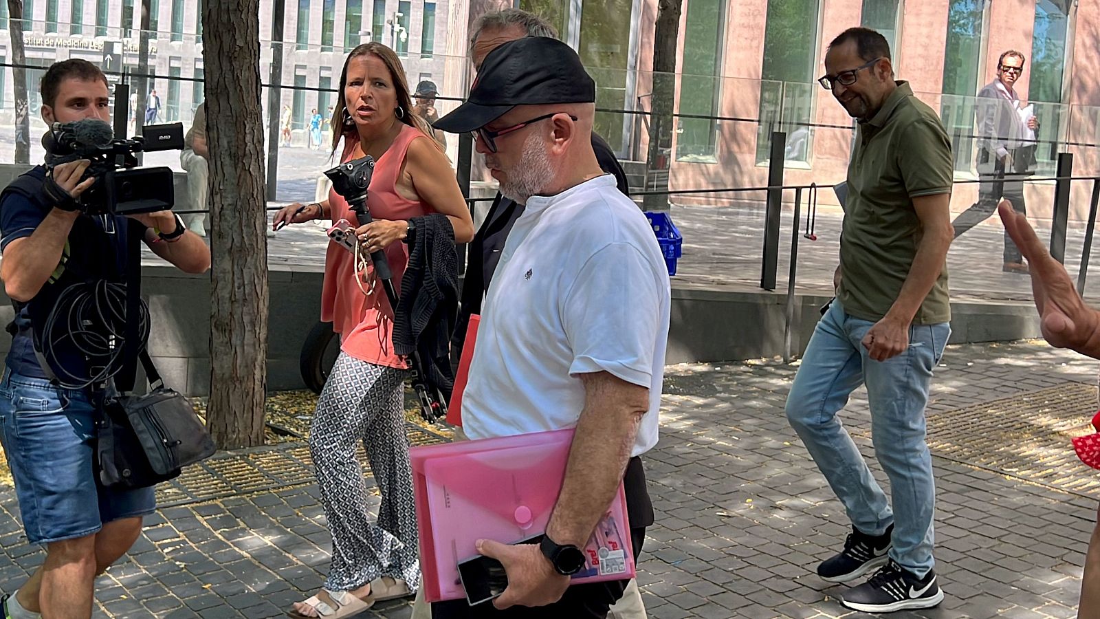 Abogado sale de un edificio rodeado de personas, entre ellas un camarógrafo y una mujer. Lleva una carpeta rosa y un teléfono móvil.