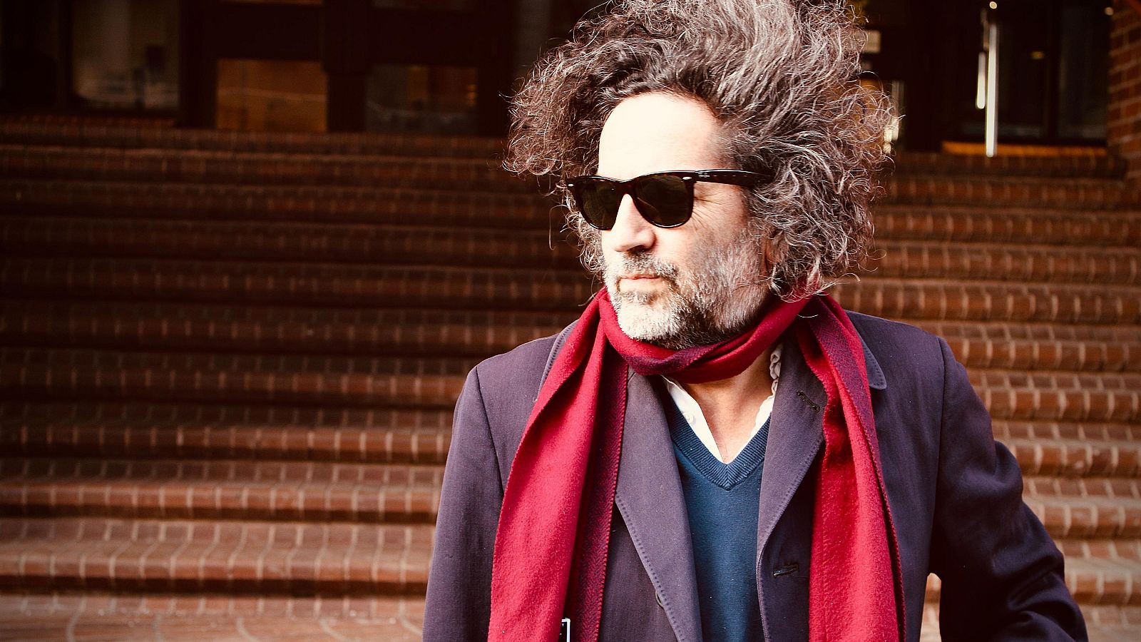 Dan Bejar, de Destroyer, en foto con gafas oscuras, bufanda roja y abrigo.  Fondo con escalera de ladrillo, iluminación cálida.
