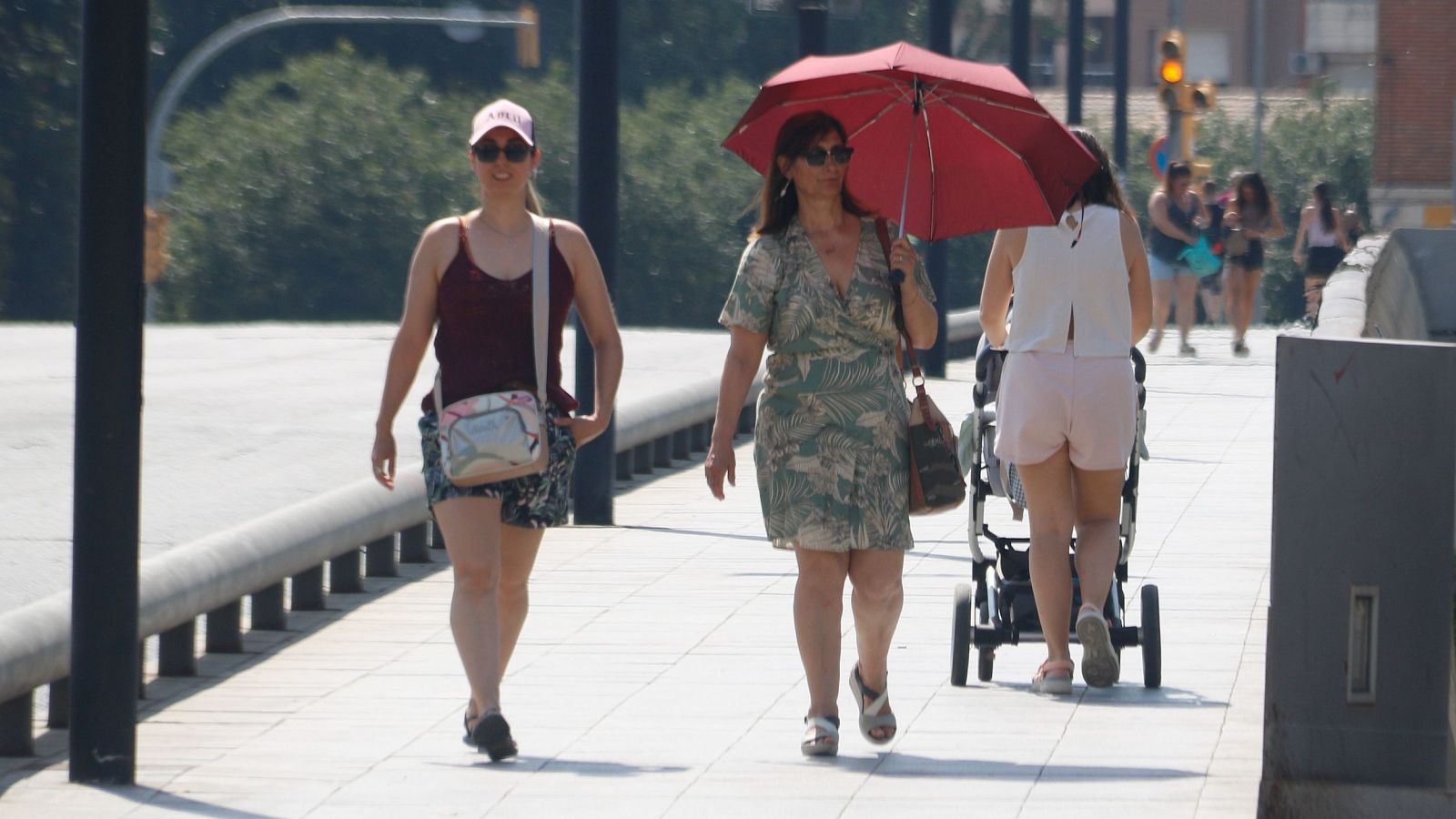 Tres mujeres, una con gorra rosa y otra con sombrilla roja, pasean por un puente soleado. Una tercera empuja un carrito de bebé en dirección contraria.
