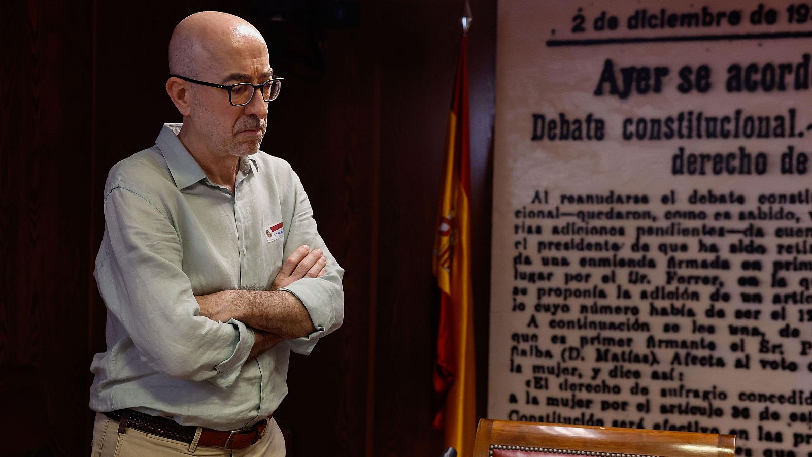 Imagen de un hombre de camisa verde claro y pantalones beige, con los brazos cruzados y expresión seria, en una sala con bandera española y un recorte de periódico enmarcado que alude al voto femenino.