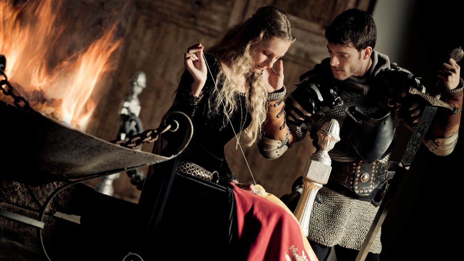 Figurines medievales: mujer cosiendo/tejiendo y hombre con espada y armadura, en un interior con chimenea.  Preparación para una escena.